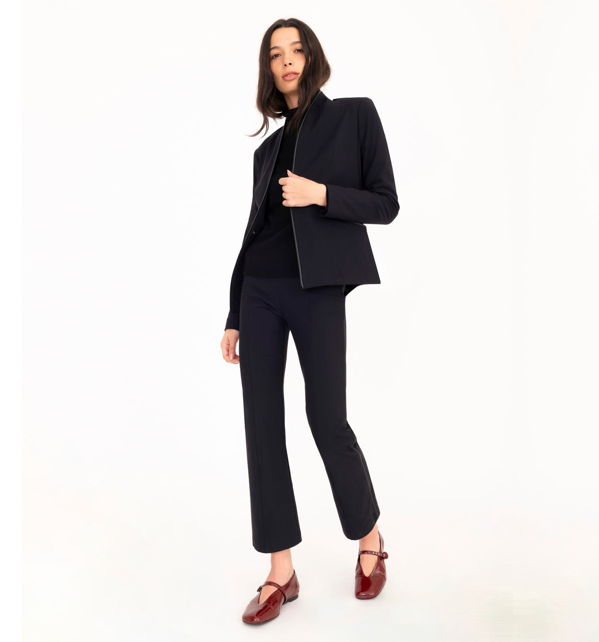 A model poses in a black tailored suit consisting of a blazer and trousers, layered over a black turtleneck sweater and paired with maroon mary jane flats, against a solid white studio background.