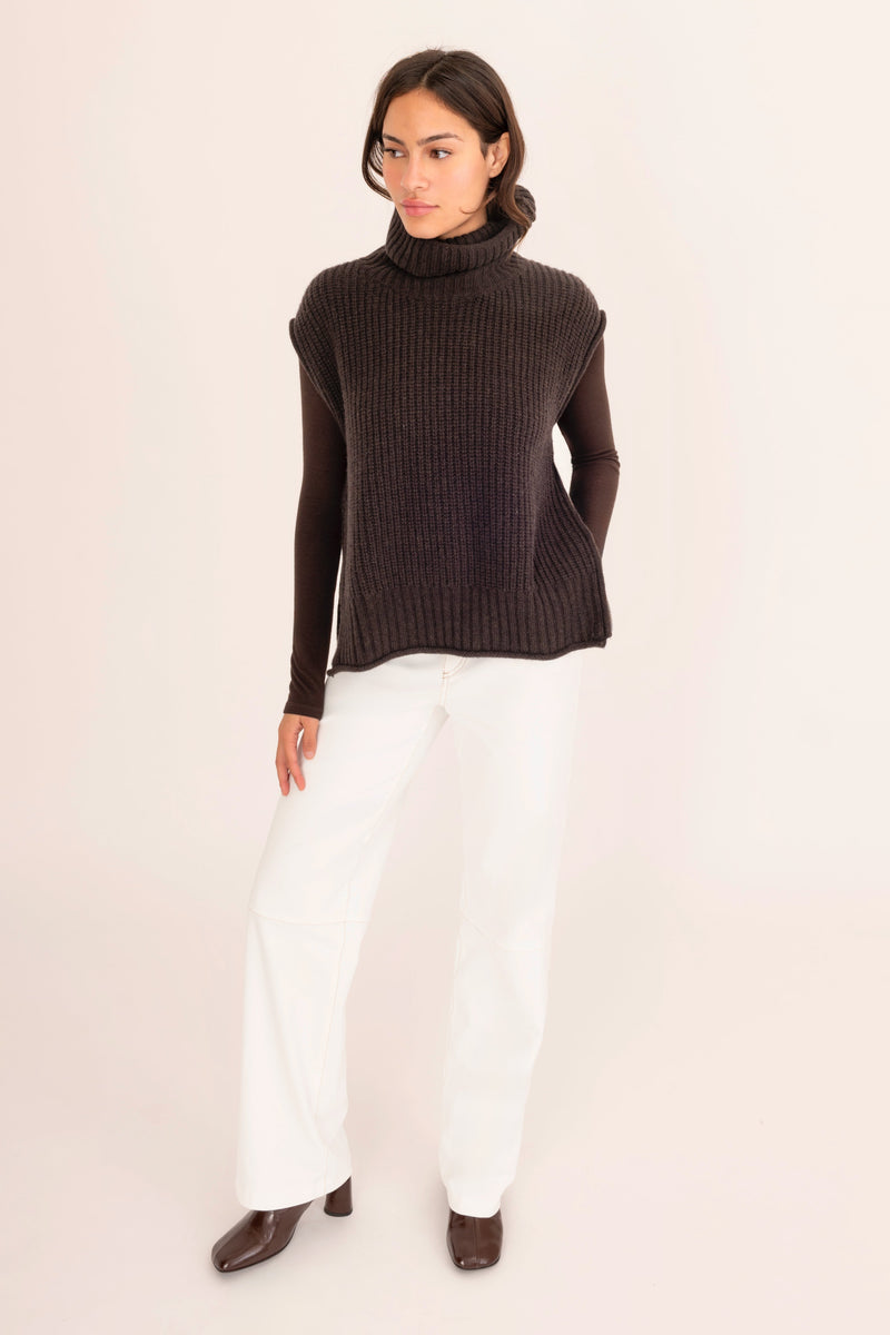 Woman wearing a dark brown turtleneck sweater and white pants on a light pink background