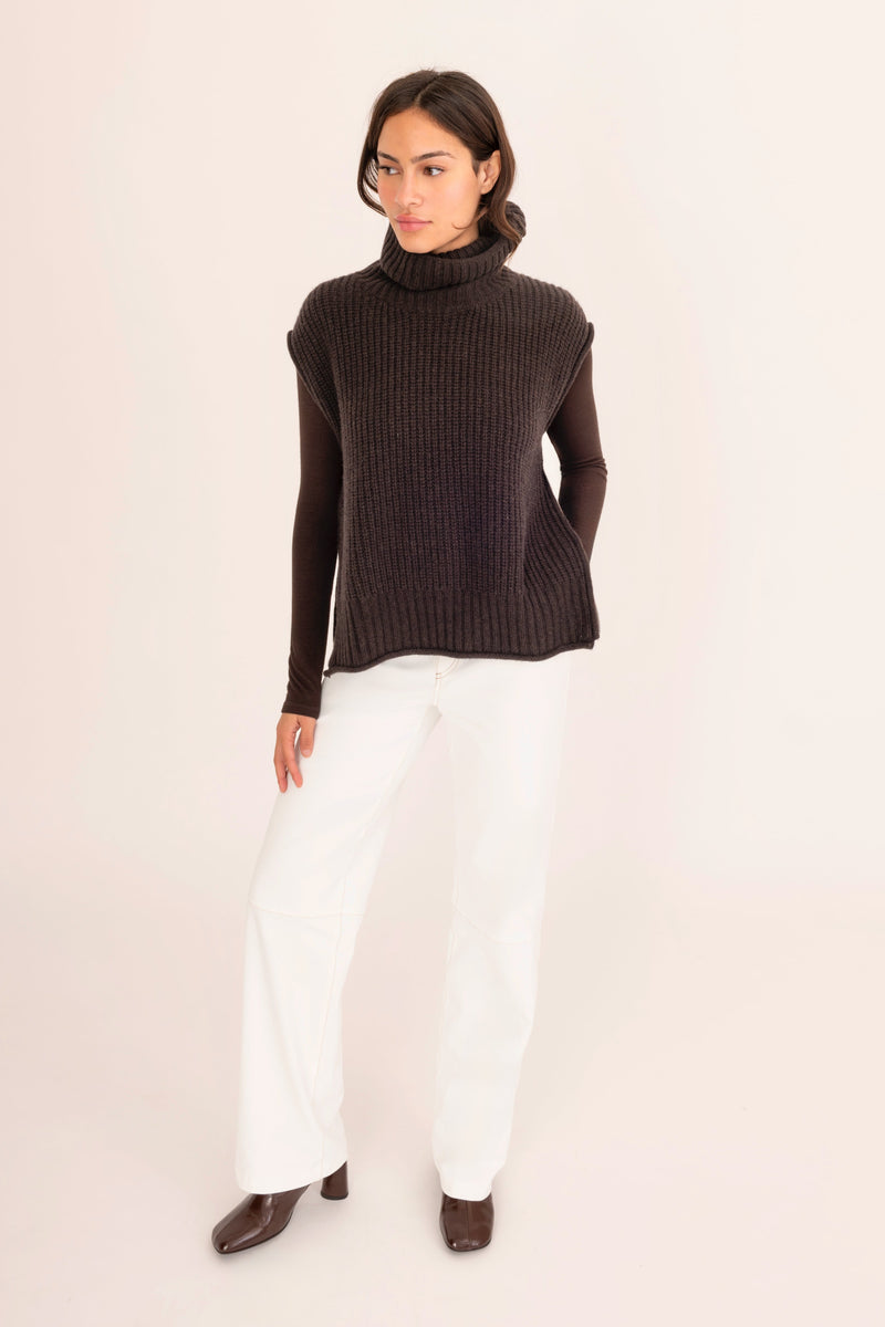 Woman wearing a dark brown turtleneck sweater and white pants on a light pink background