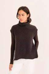 Woman wearing a dark brown turtleneck sweater and white pants on a light pink background