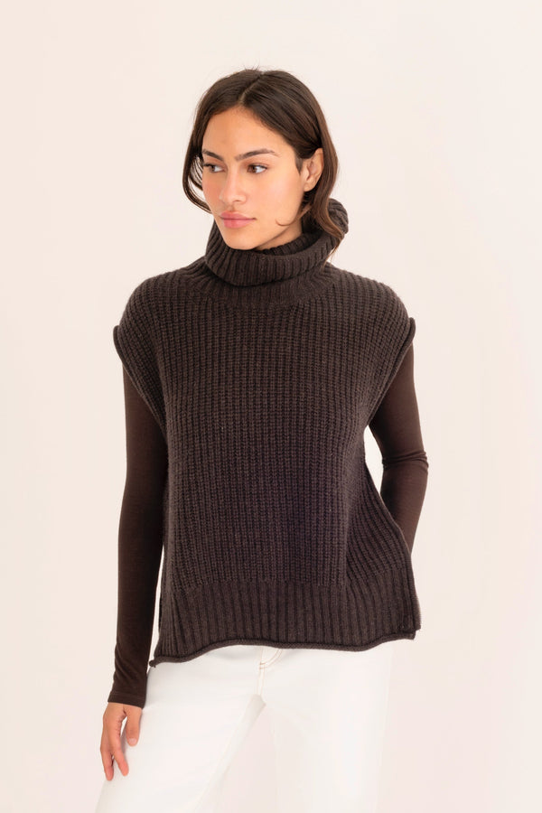 Woman wearing a dark brown turtleneck sweater and white pants on a light pink background