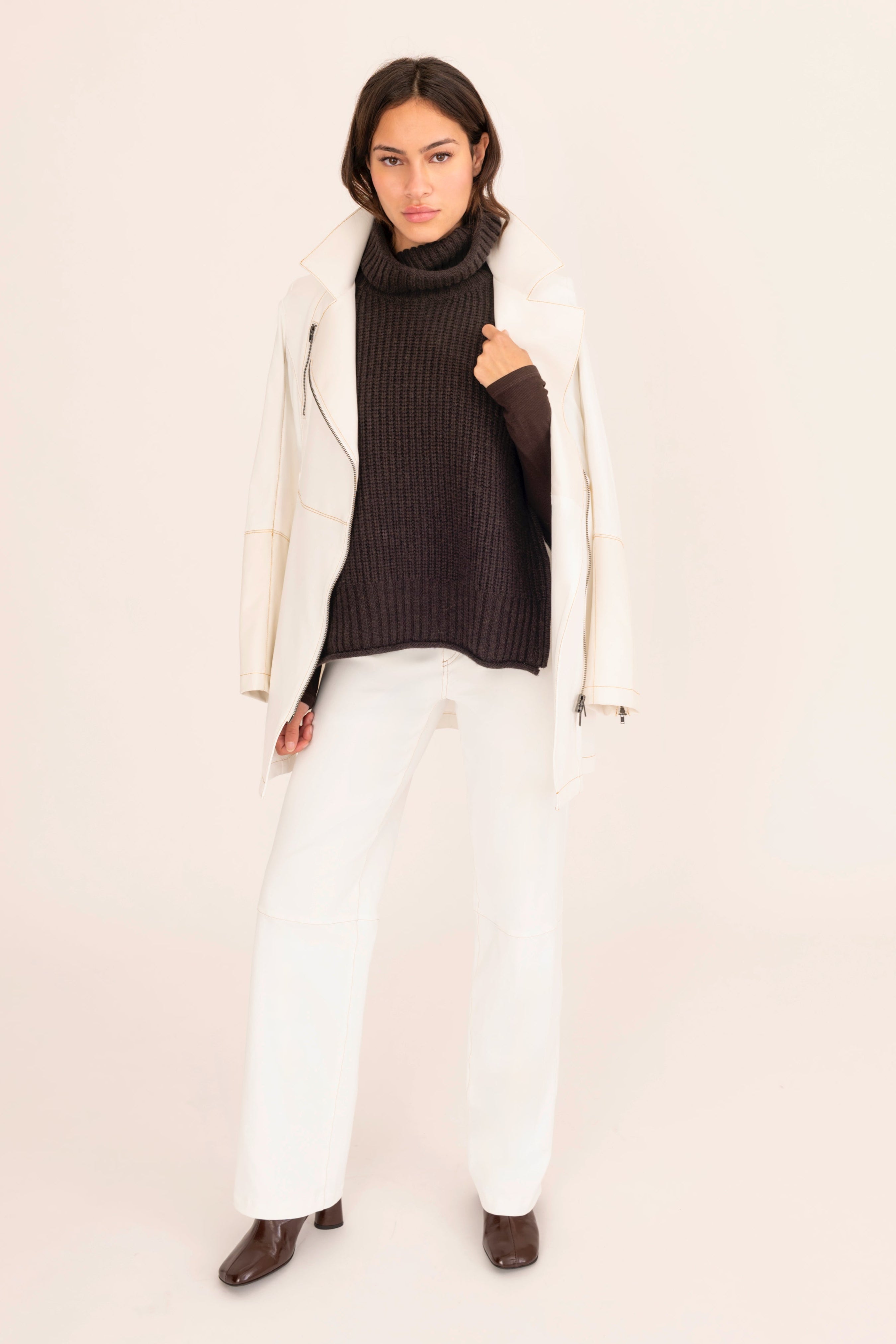 Person wearing a brown turtleneck, white jacket, and white pants on a beige background