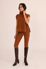 Woman wearing a brown sleeveless sweater and leggings with knee-high boots on a beige background