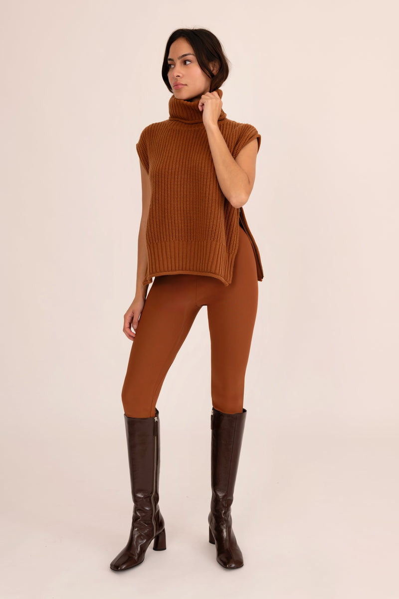 Woman wearing a brown sleeveless sweater and leggings with knee-high boots on a beige background