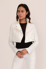 Woman wearing a white jacket over a black top and white pants on a light pink background