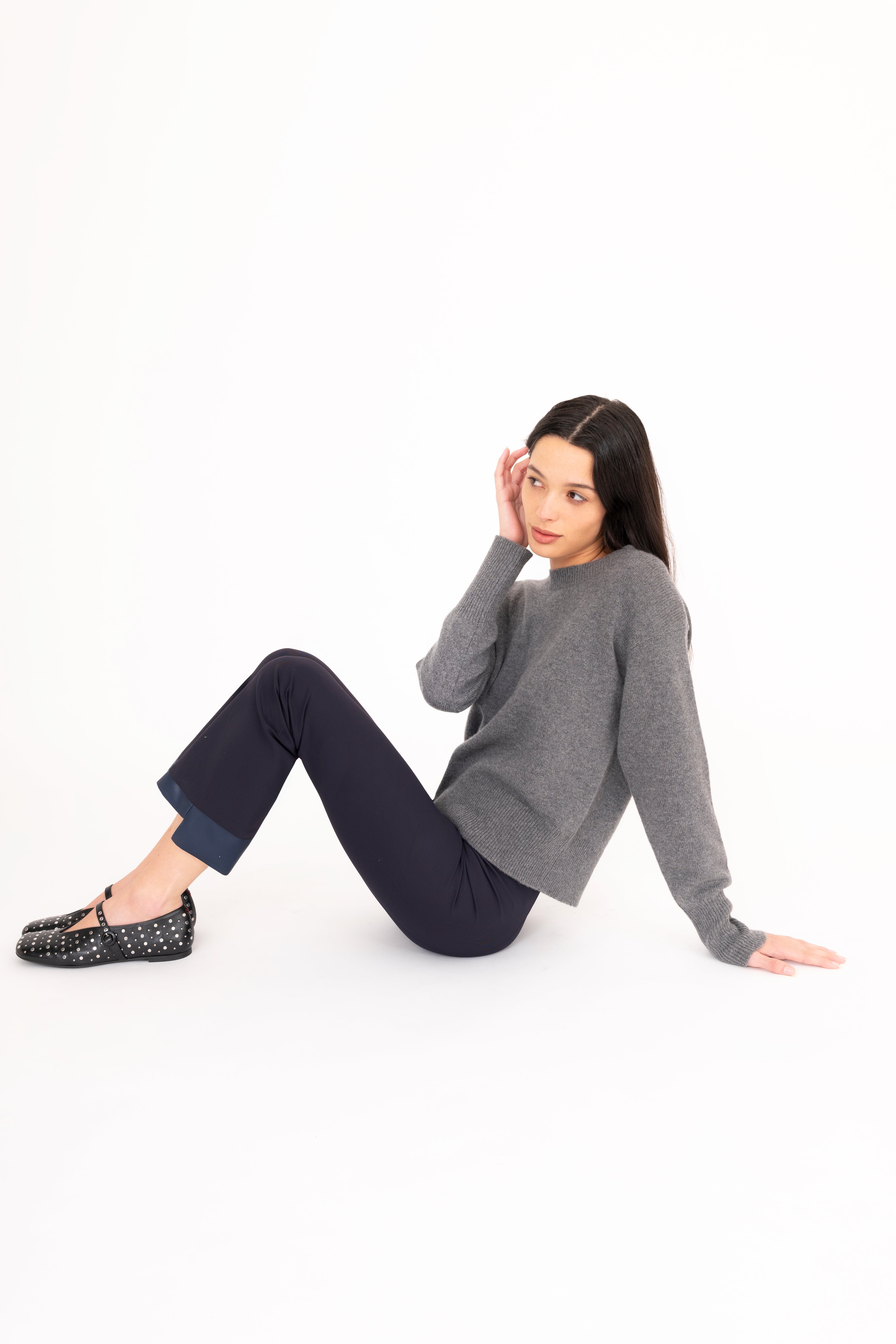 Woman sitting on the floor wearing a gray sweater and dark pants against a white background