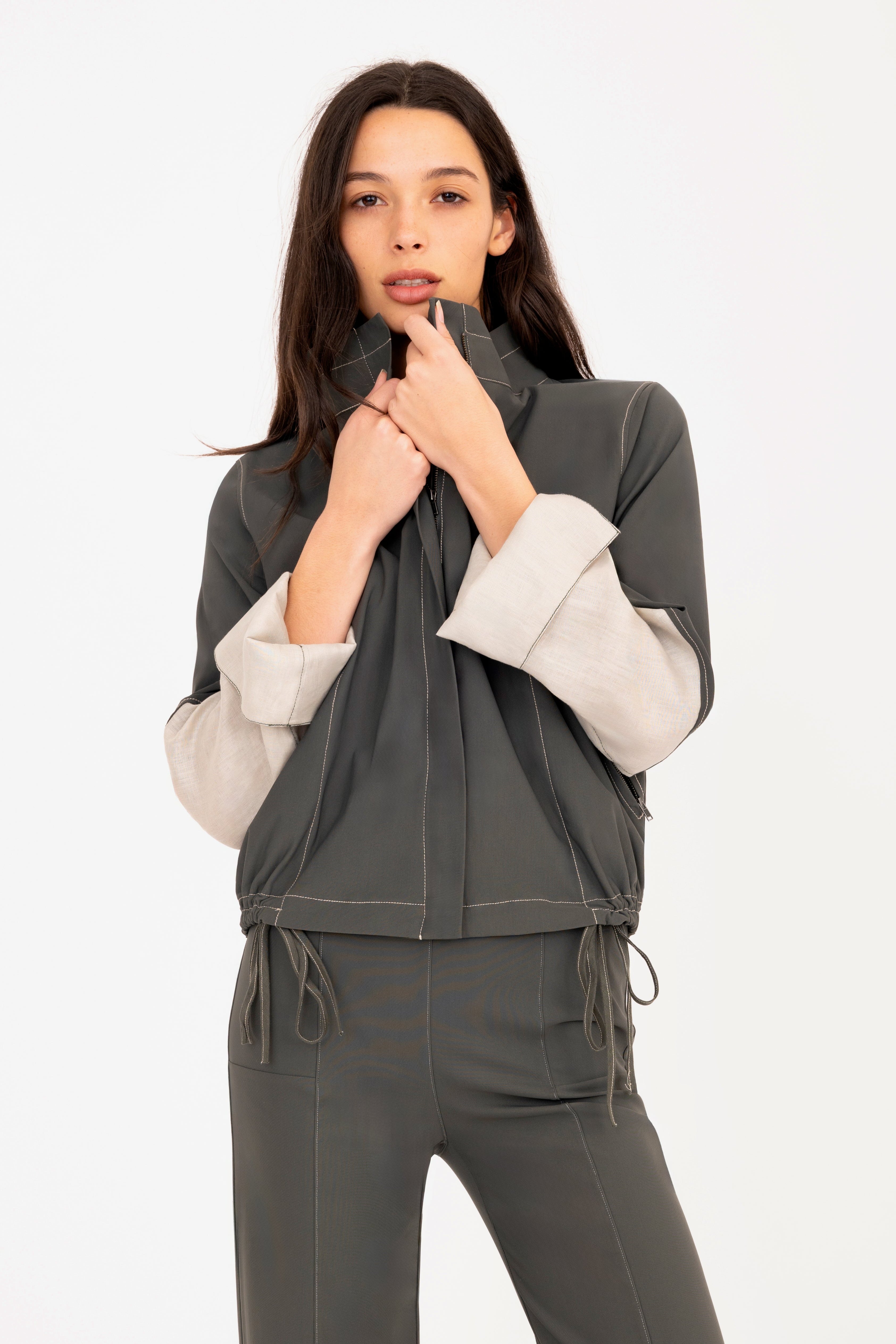 A woman modeling a dark green zip-up jacket with contrasting light beige sleeves and matching dark green drawstring pants, set against a plain white studio background.