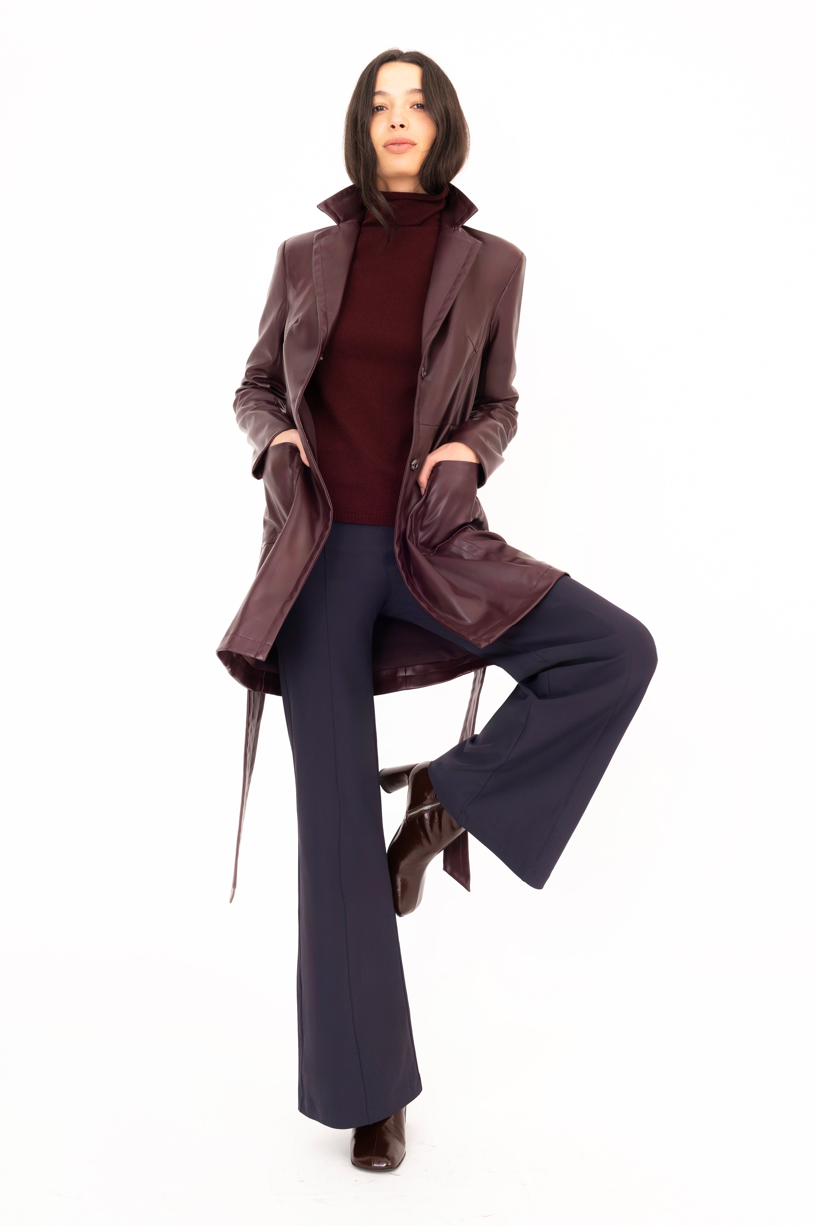 Woman wearing a brown coat and dark pants sitting on a chair against a white background