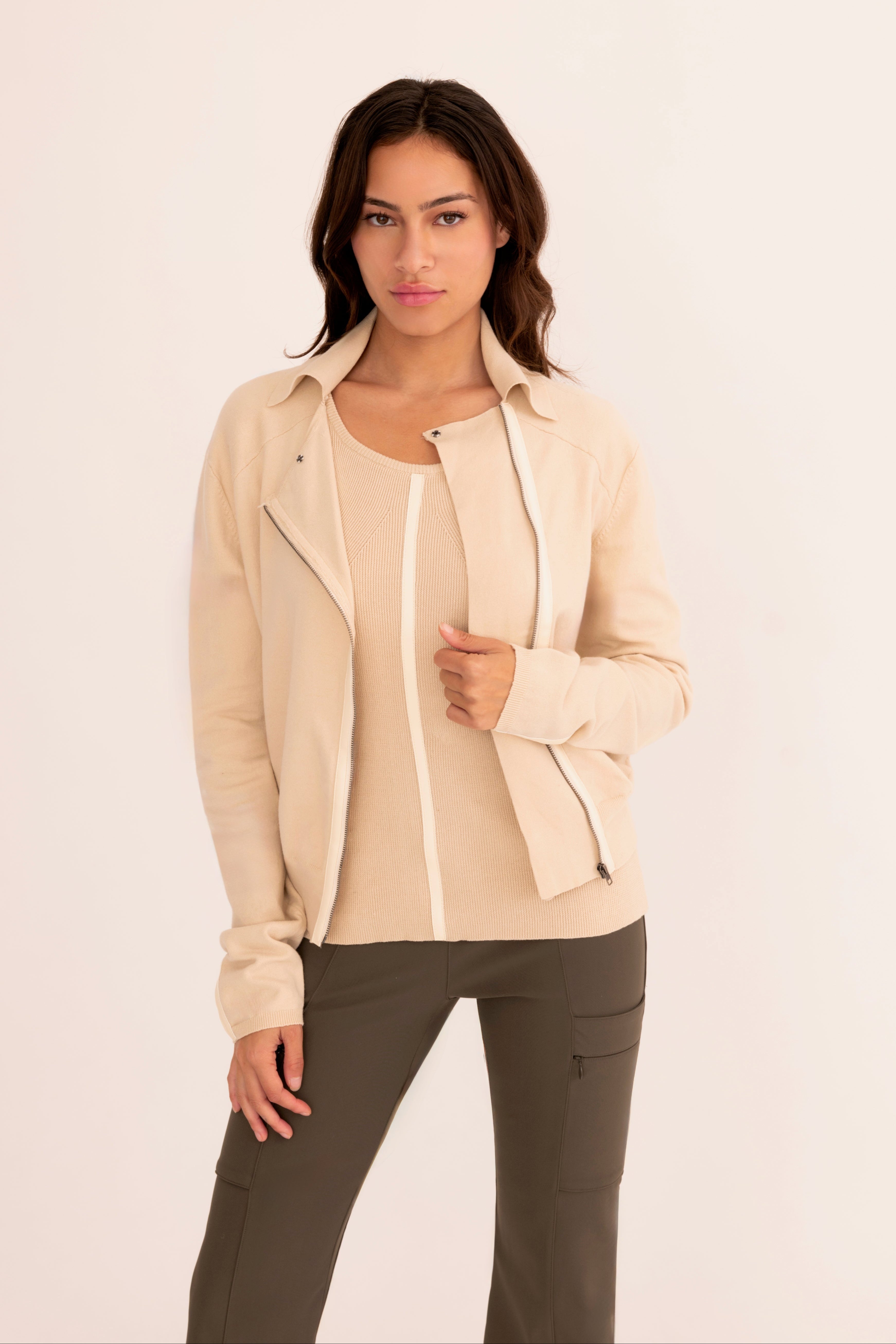A woman modeling a light beige zip-up jacket over a matching beige top and dark gray pants, standing against a plain off-white studio background.