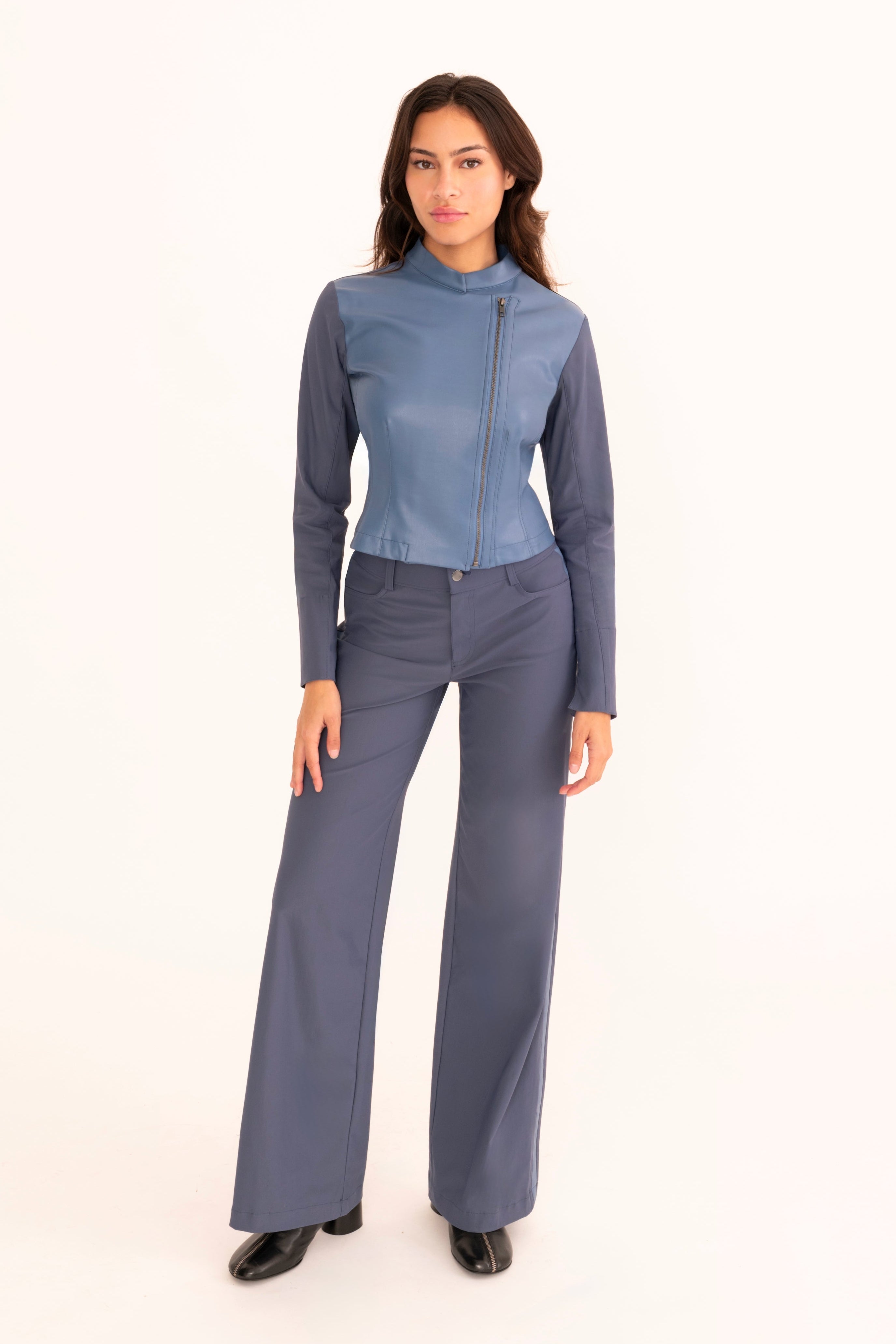 A woman modeling a blue faux-leather jacket and wide-leg black trousers, standing against a solid white studio background.