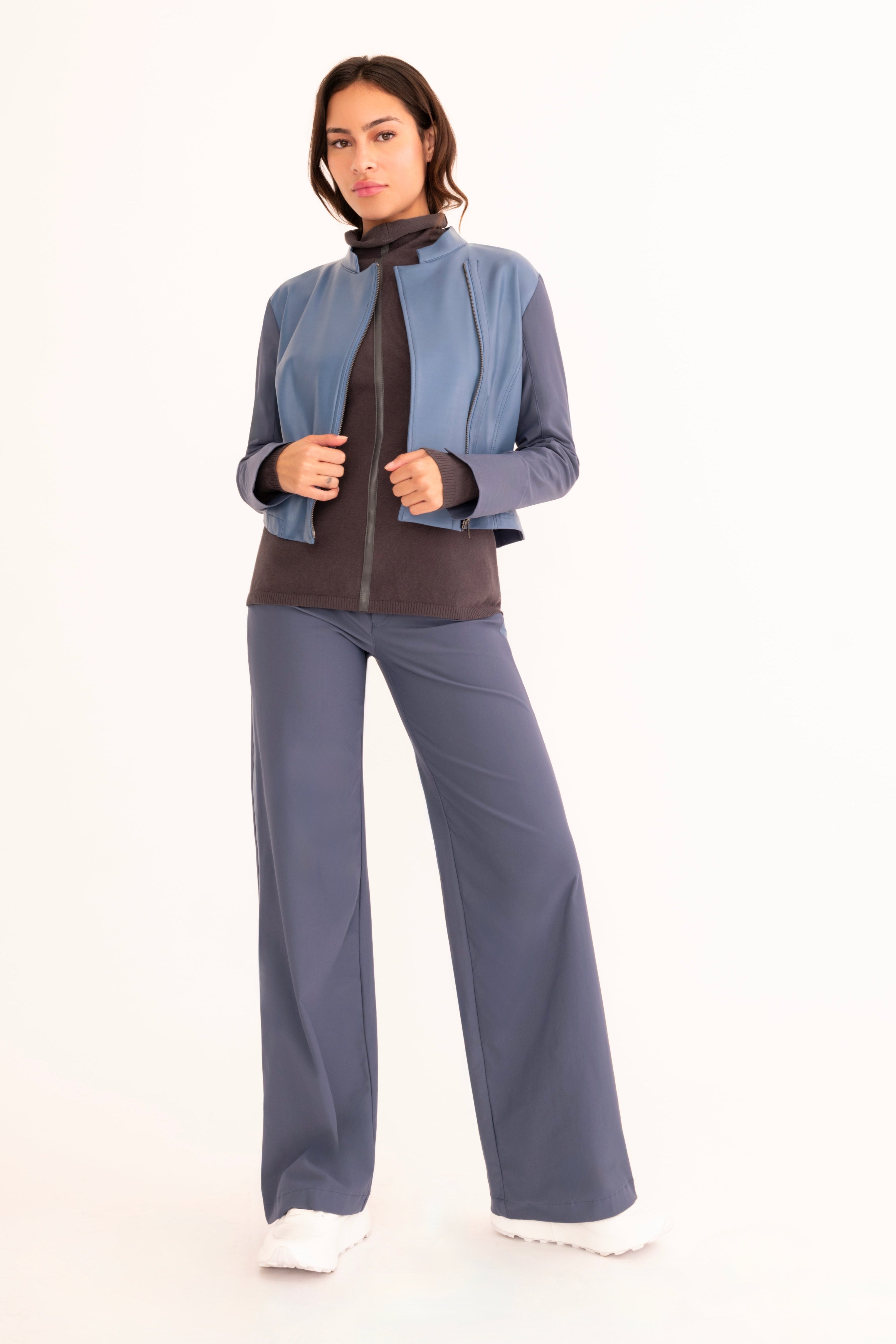 A woman modeling a matching two-piece tracksuit featuring a blue cropped, zip-front jacket  paired with matching wide-leg trousers in a blue with a gray mock-neck sweater, on a solid white studio background.