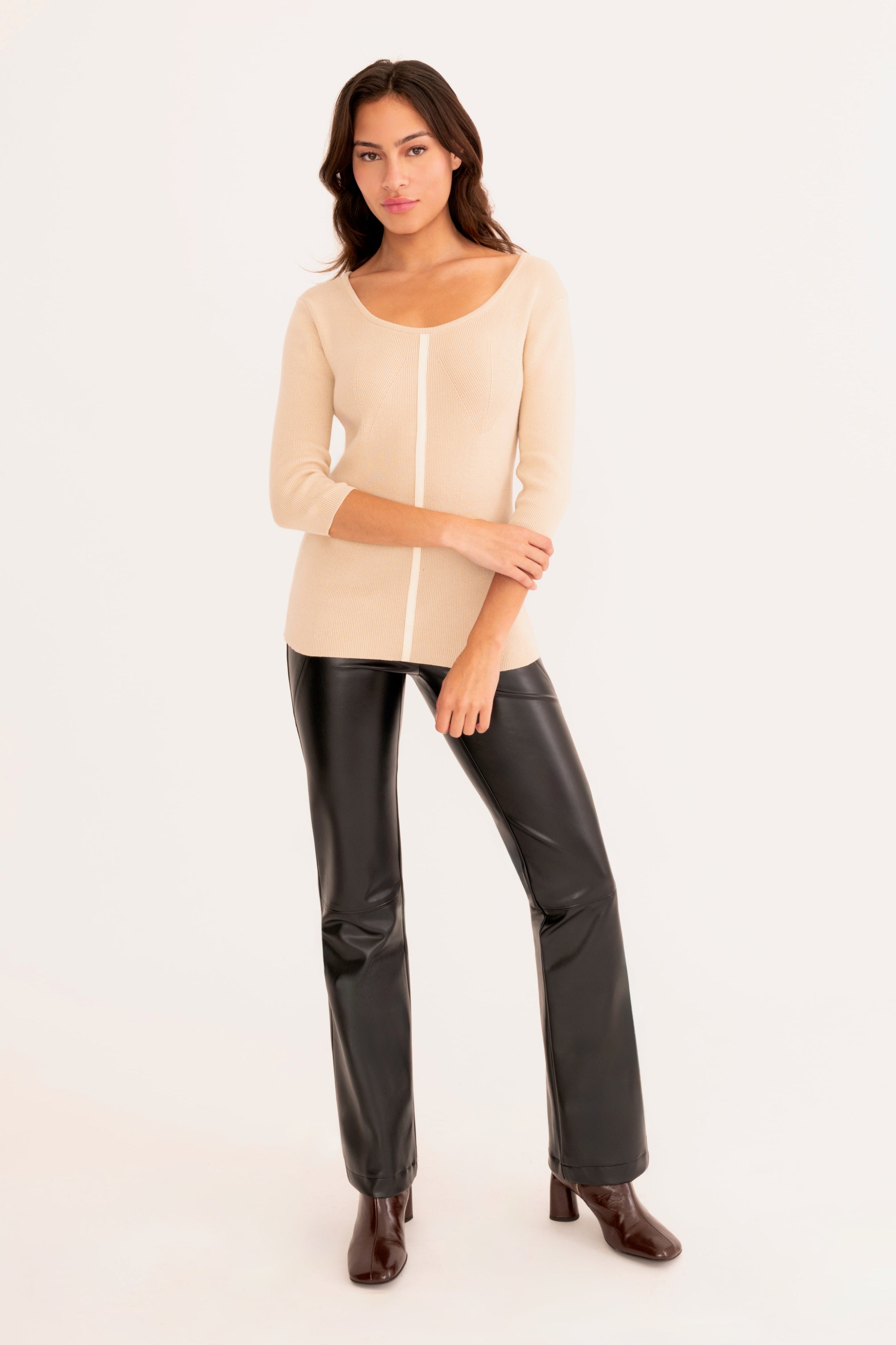 A woman modeling a beige knit top  and black faux-leather pants against a plain white studio background.