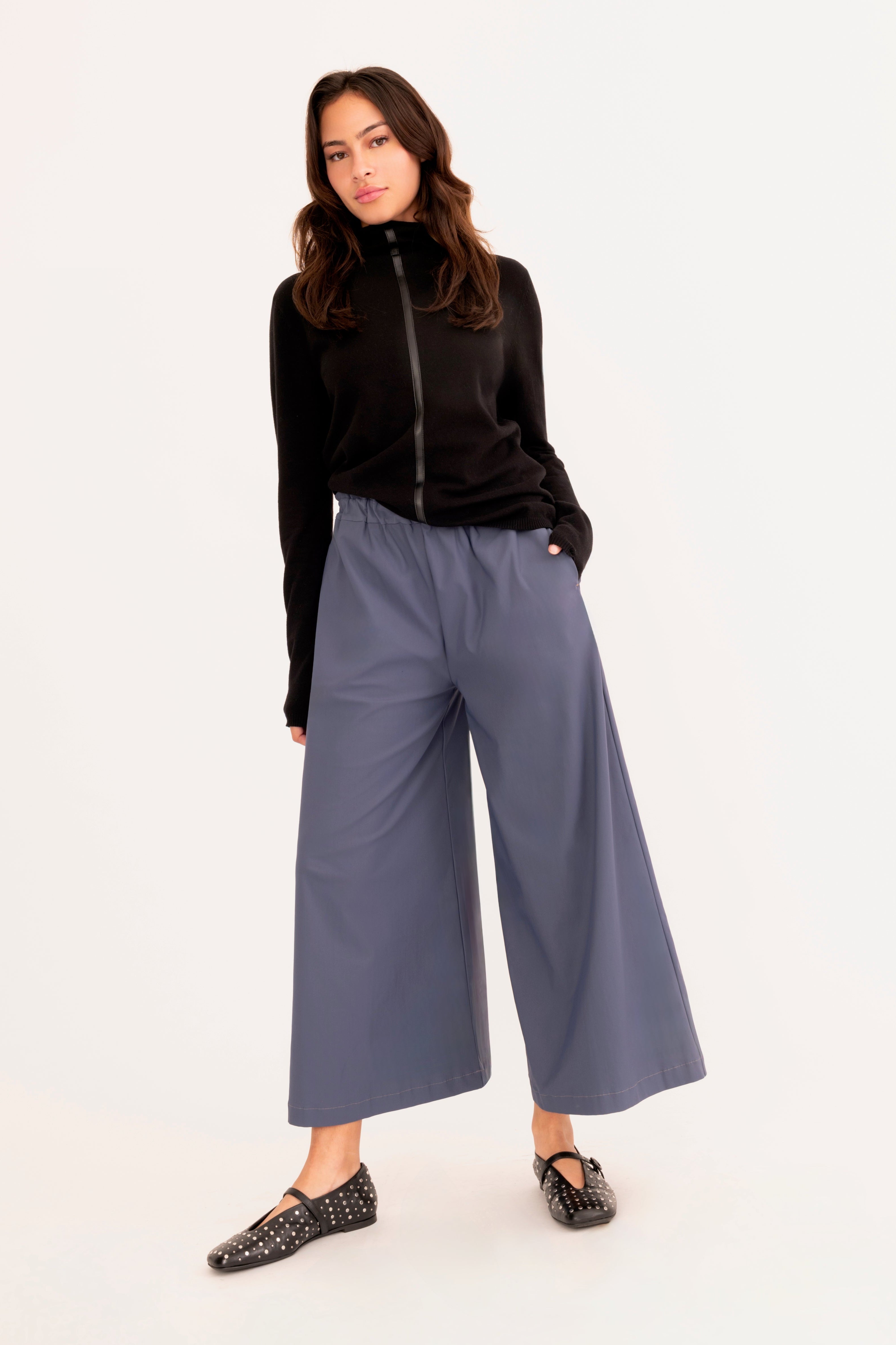 A woman modeling a pair of wide-leg cropped pants in slate blue, paired with a black mock-neck long-sleeve top and black studded loafers, against a plain white studio background.