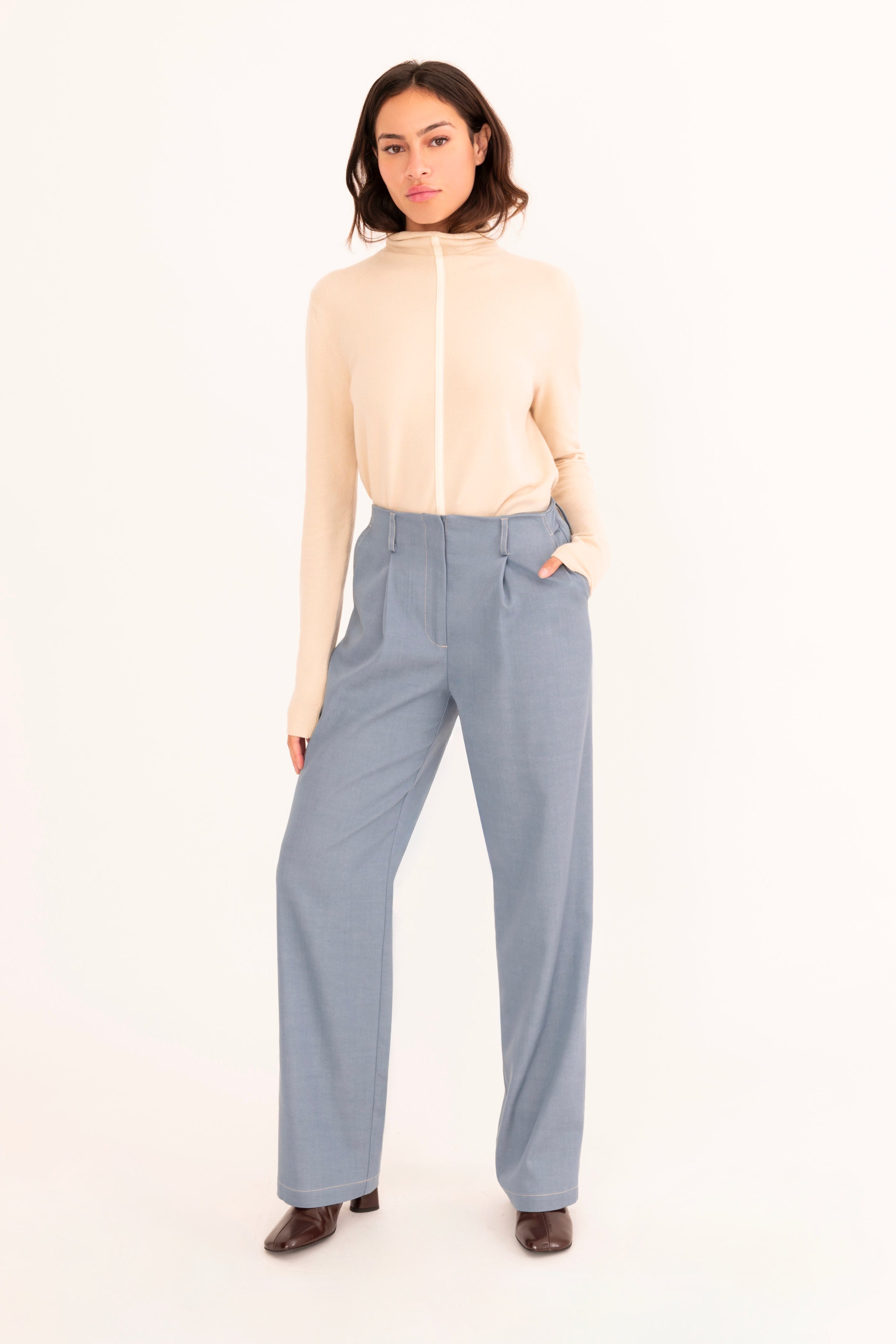 A woman stands against a plain white studio background wearing a light beige mock-neck knit top and high-waisted, wide-leg blue trousers.