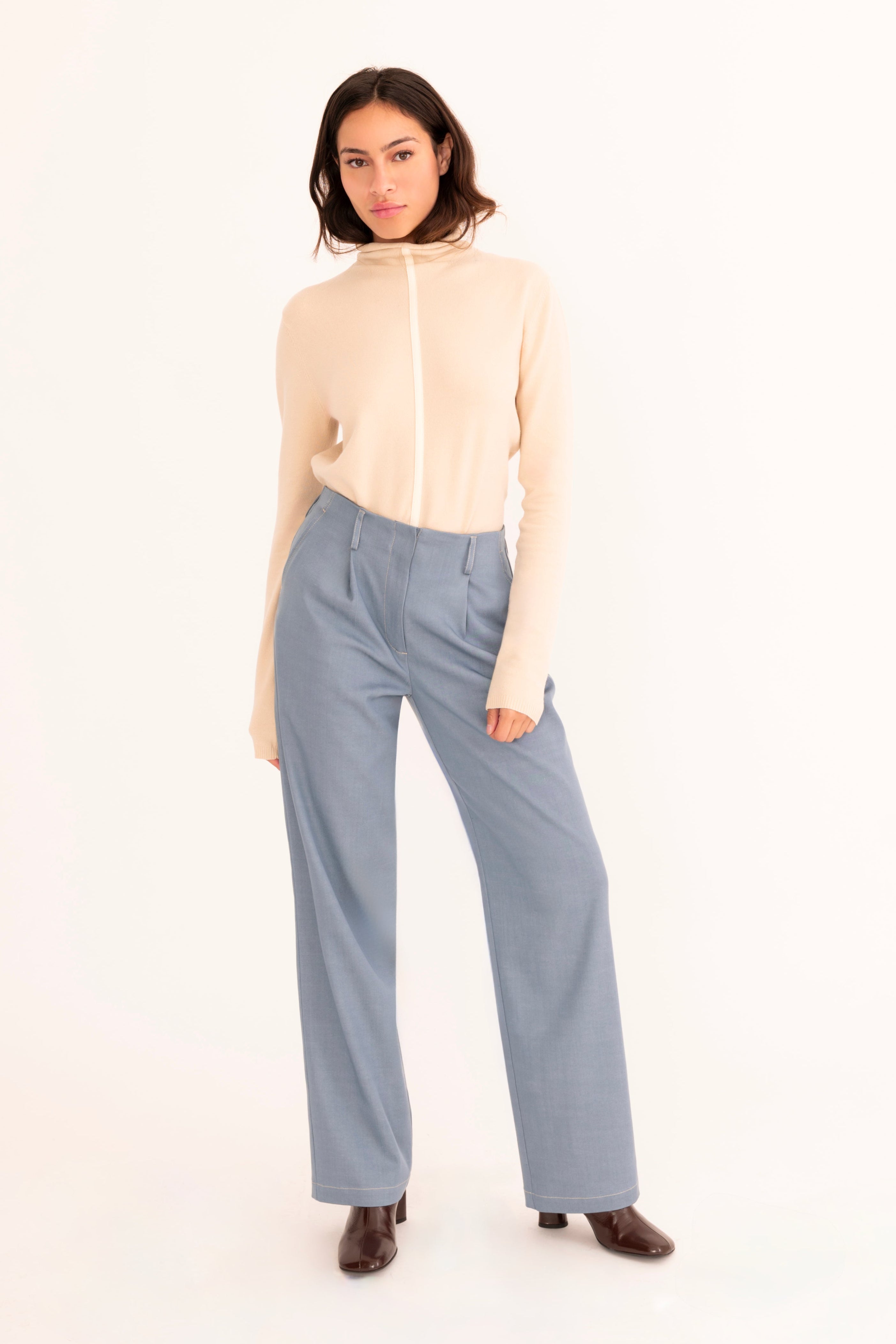 A woman stands against a plain white studio background wearing a light beige mock-neck knit top and high-waisted, wide-leg blue trousers.