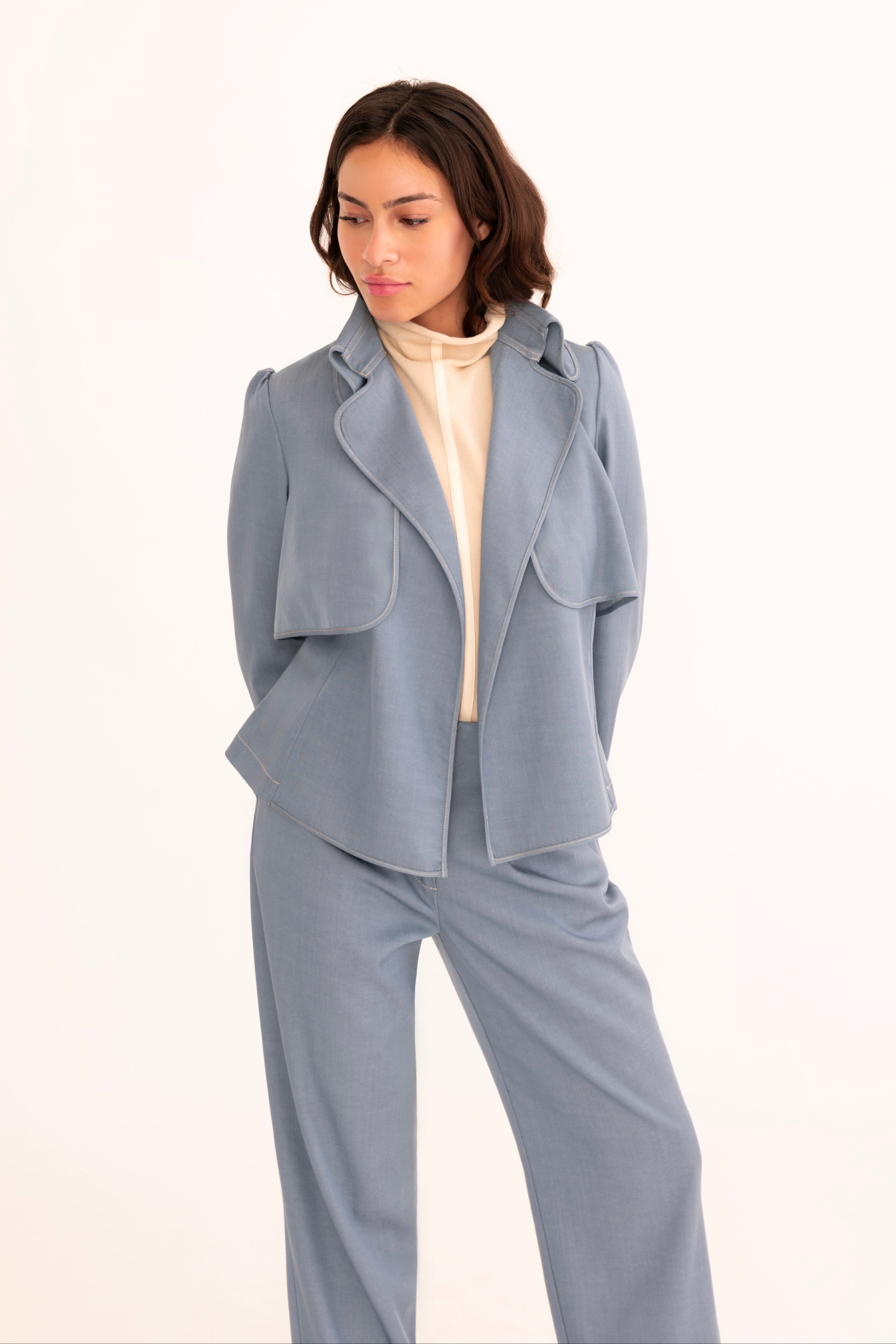 A model wearing a light blue tailored jacket and matching wide-leg trousers over a cream mock-neck top, standing against a plain white studio background.