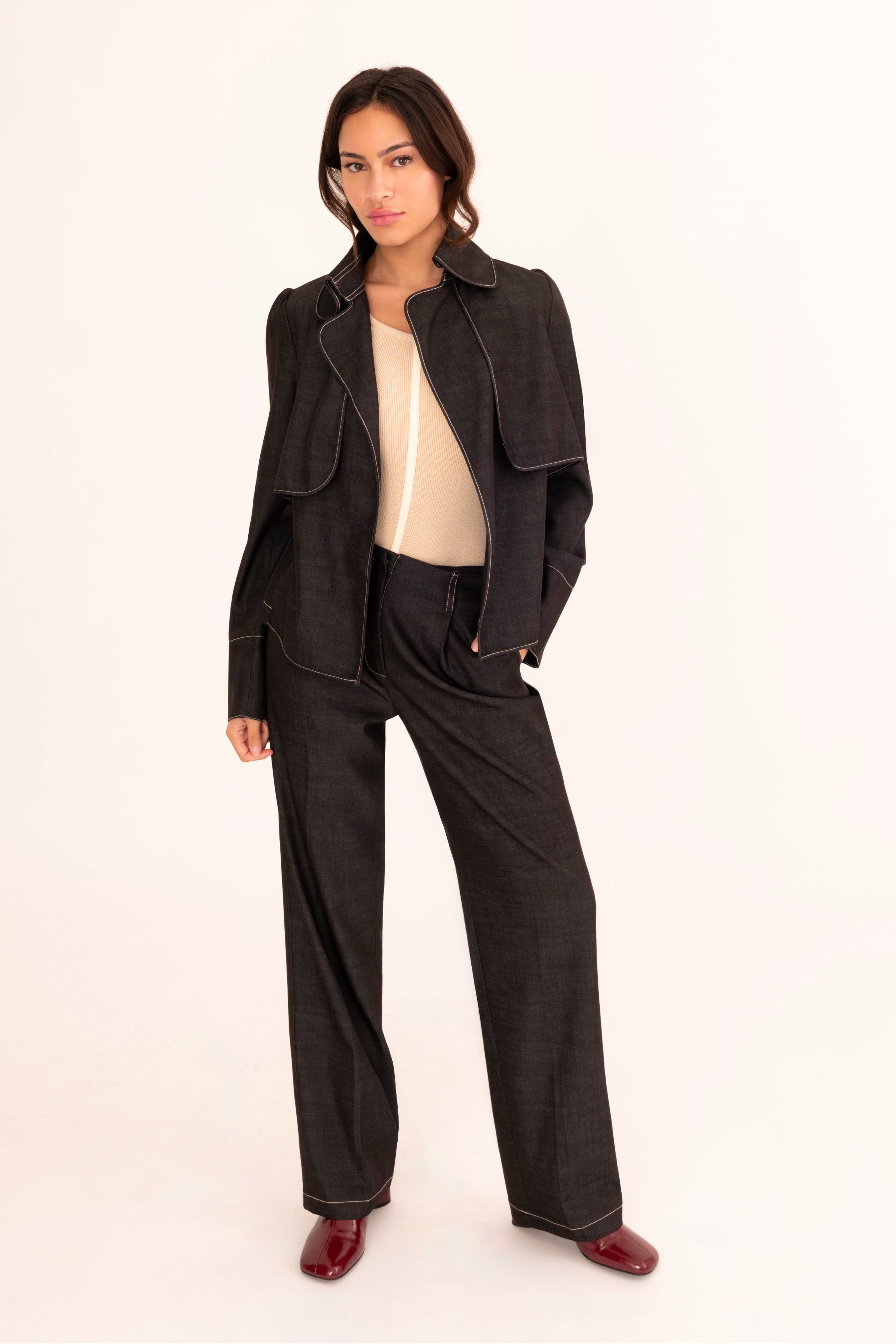 A model stands against a plain white studio background wearing a black cropped jacket, black wide-leg trousers, a cream top, and dark red shoes.