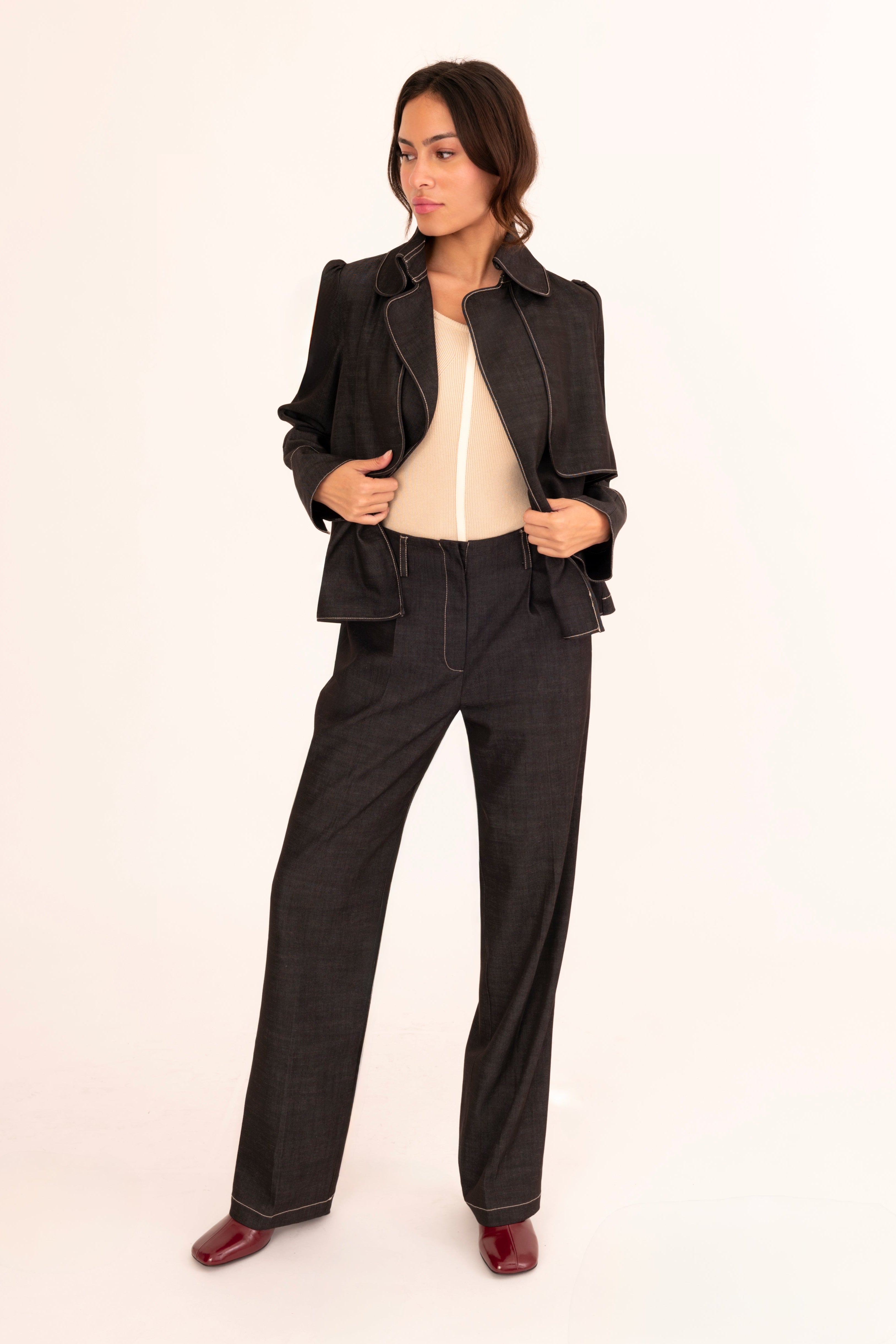 A model stands against a plain white studio background wearing a black cropped jacket, black wide-leg trousers, a cream top, and dark red shoes.