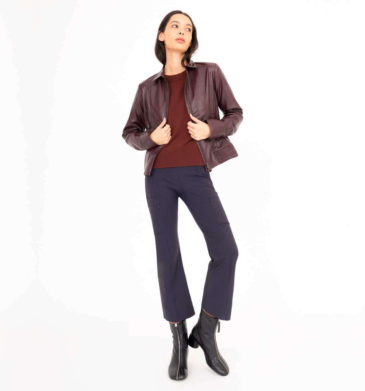 A woman modeling a dark brown leather jacket over a burgundy top, navy blue pants, and black ankle boots, standing against a solid white background.