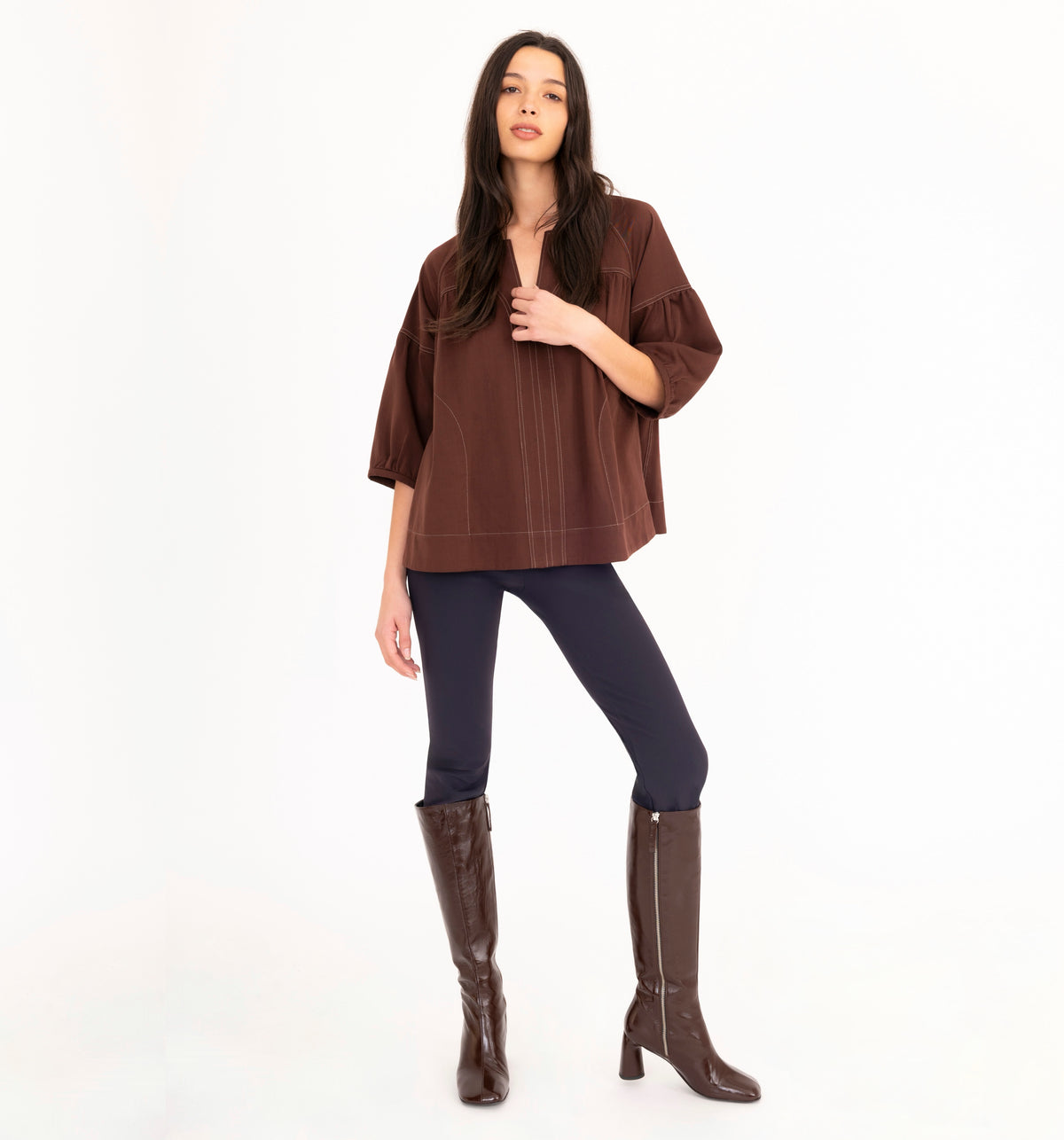 A woman modeling a brown 3/4-sleeve top with vertical seam details, paired with dark navy pants and brown knee-high boots, against a plain white studio background.