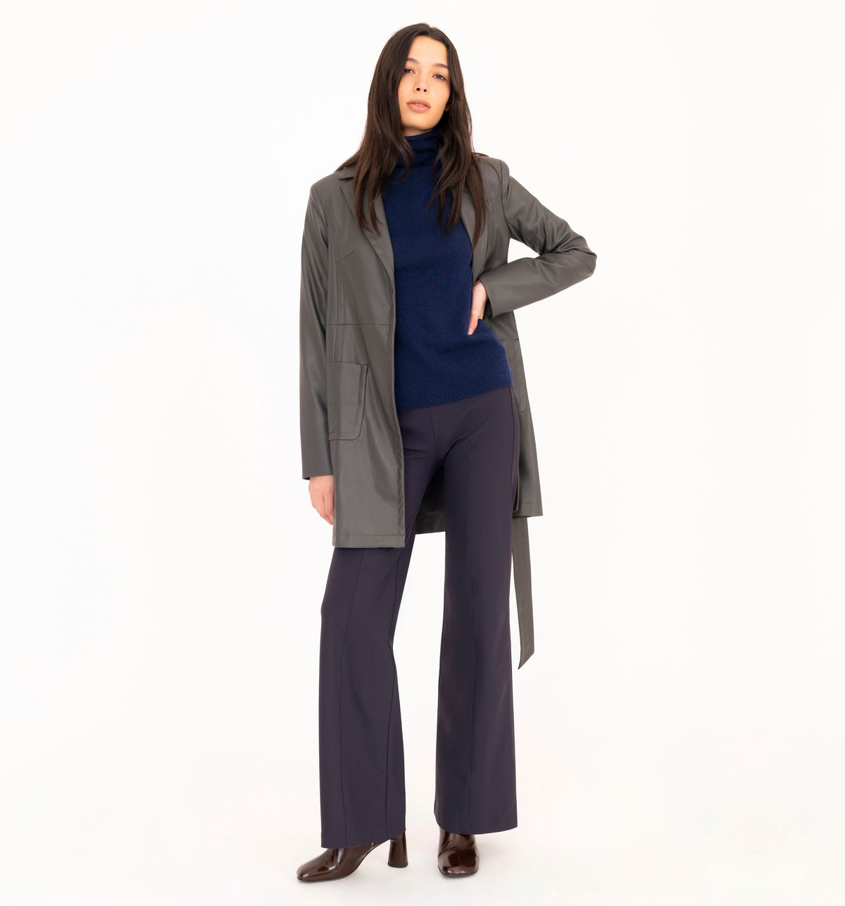 A model wearing a gray vegan leather trench coat over a blue cashmere turtleneck, navy wide-leg trousers, and brown leather boots against a solid white background.