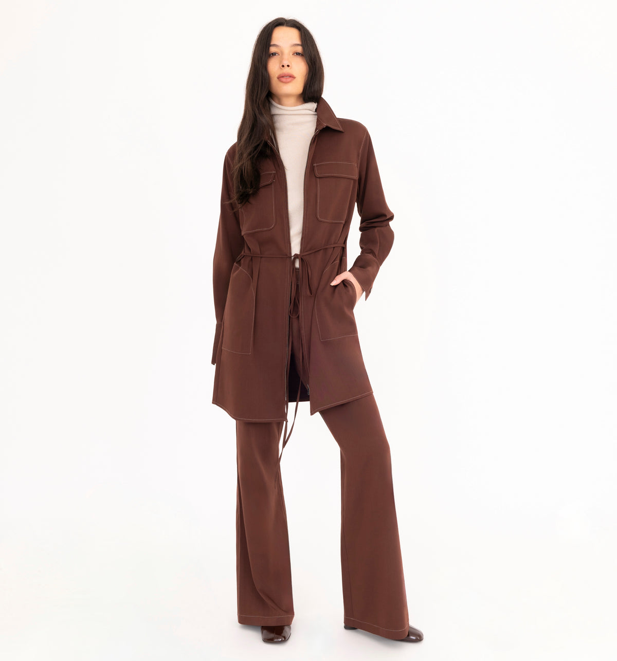 A woman modeling a brown denim shirt dress worn as a jacket with matching flared denim trousers, and a beige cashmere mock-neck sweater, paired with dark brown leather boots, against a plain white studio background.