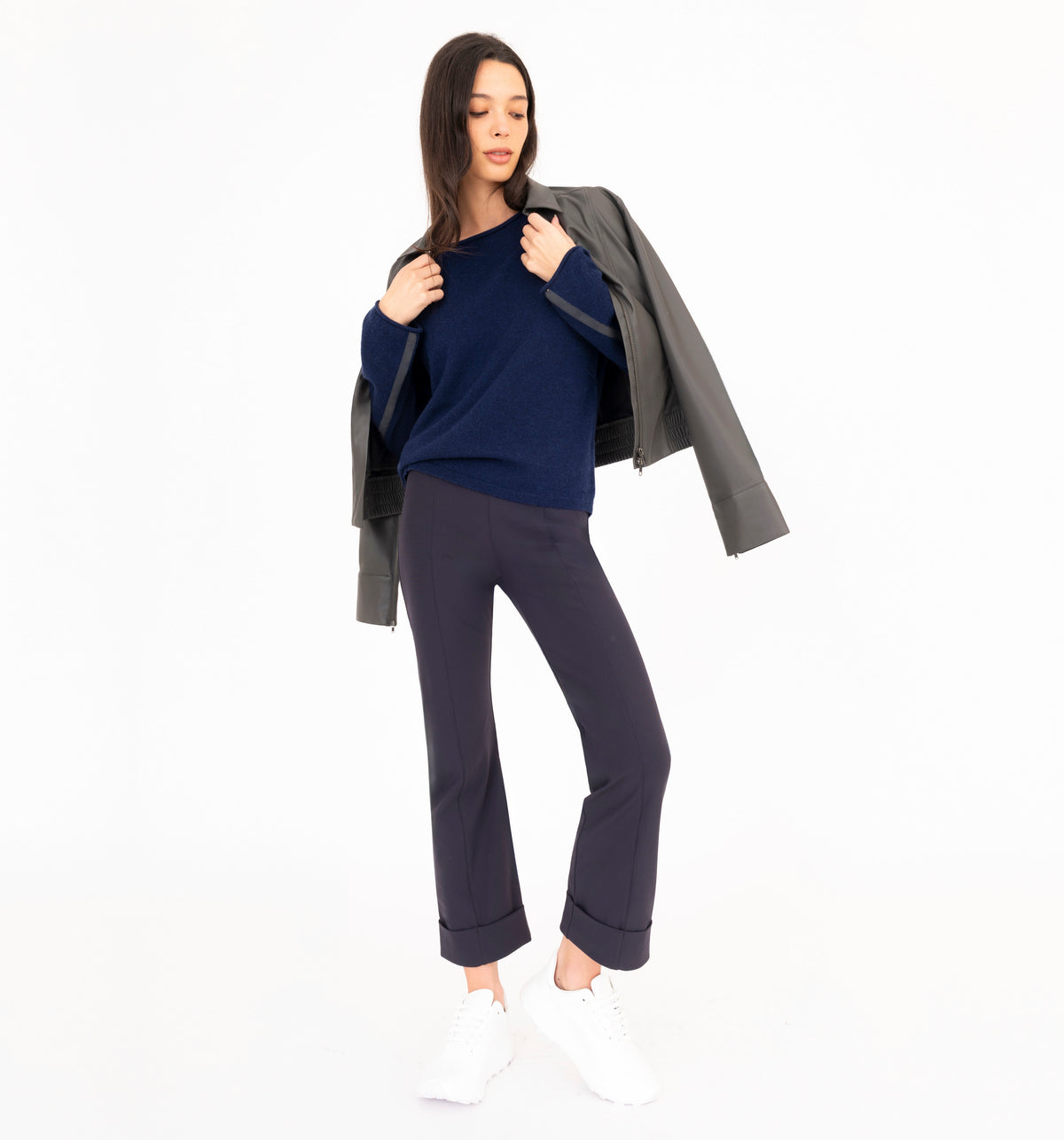 A woman modeling a navy blue cashmere sweater, navy blue cropped pants, white sneakers, and holding a gray vegan leather jacket against her shoulders, shot against a plain white studio background.