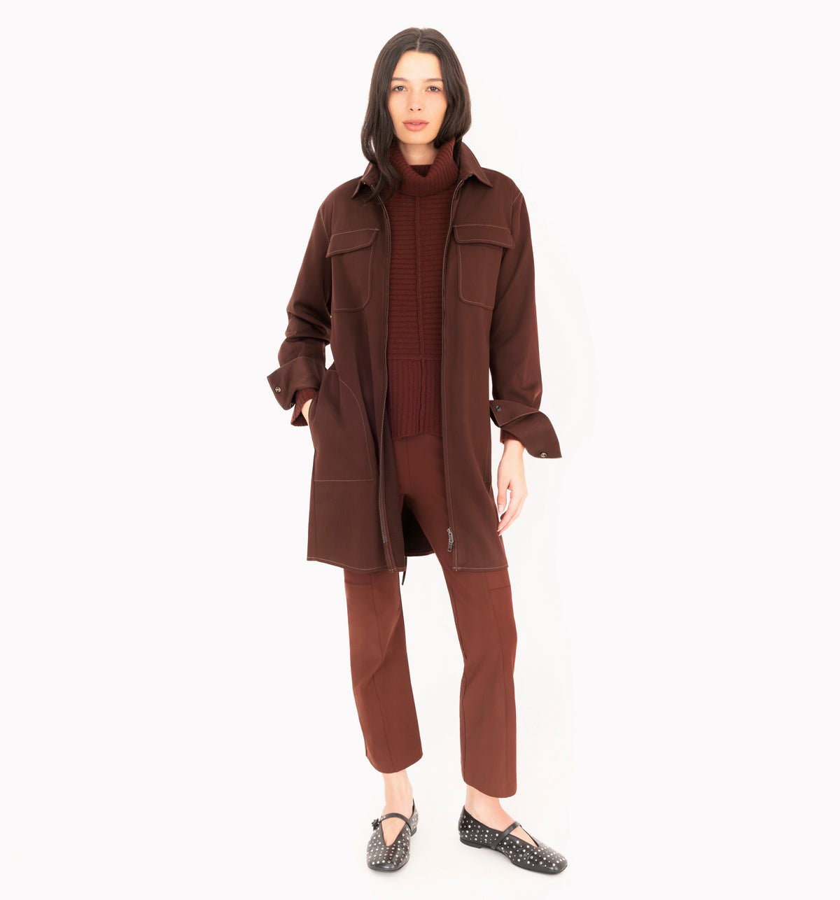 A model stands against a solid white background wearing a matching dark brown three-piece clothing set consisting of a long-sleeve button-up jacket, a ribbed turtleneck sweater, and cropped trousers, paired with black patterned flat shoes.