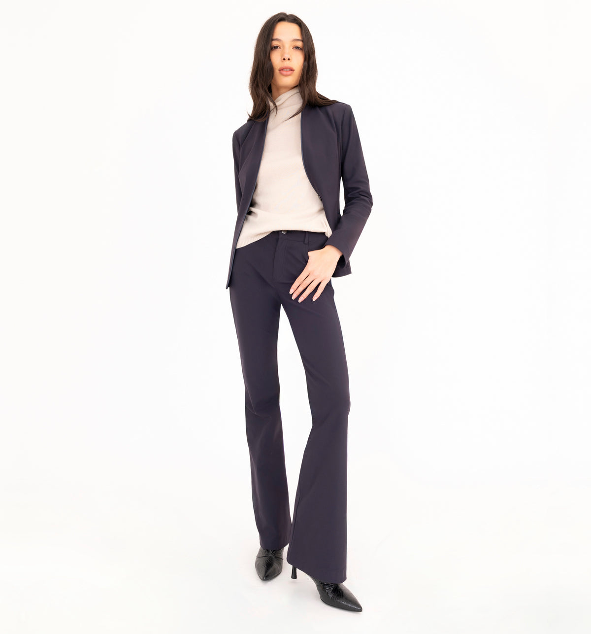A woman modeling a dark navy blue pantsuit, consisting of a tailored blazer and matching flare jean pants, styled with a beige cashmere mock neck top and black pointed-toe heels against a solid white studio background.