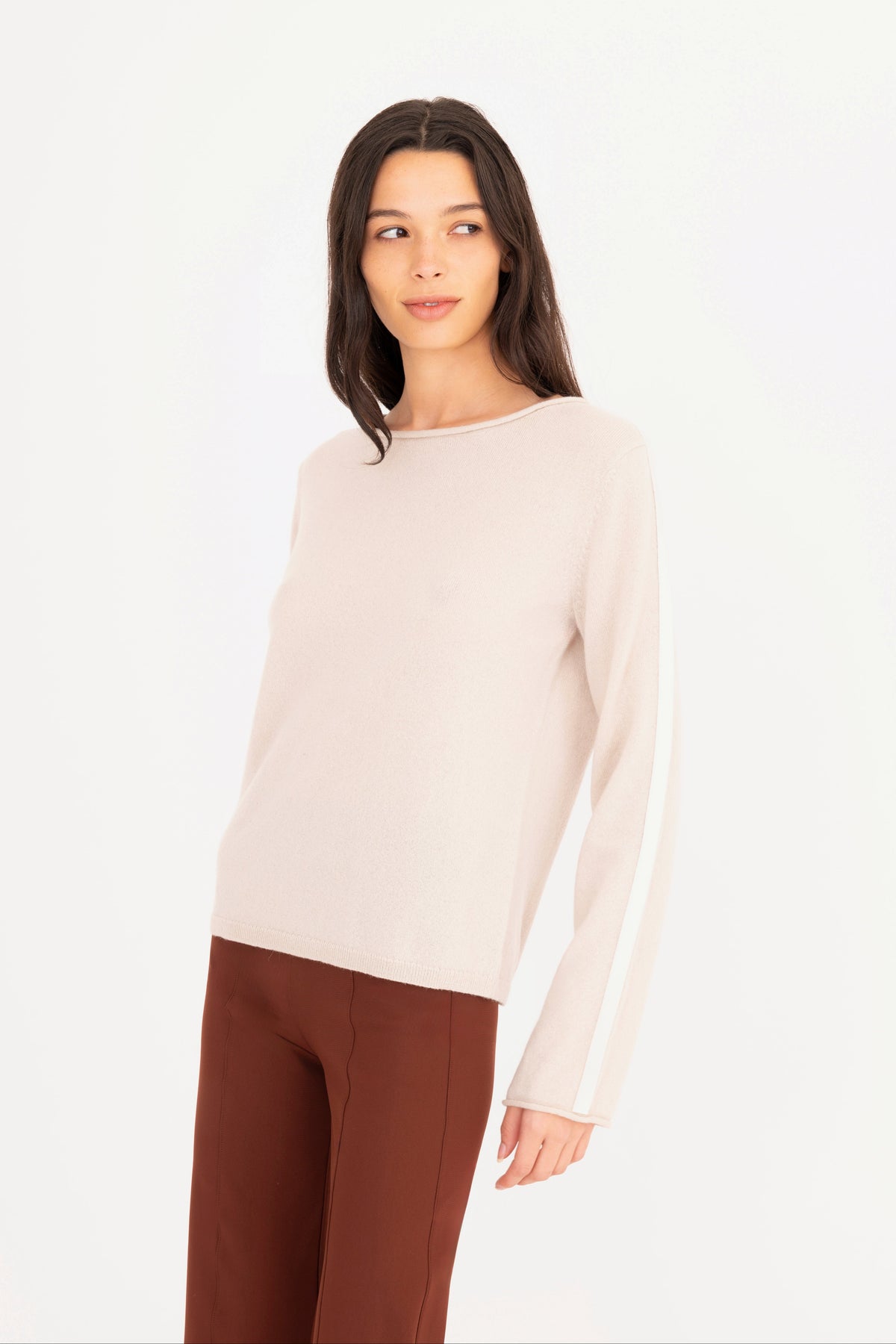 A woman modeling a beige long-sleeve knit sweater, paired with brown pants, against a plain white studio background.