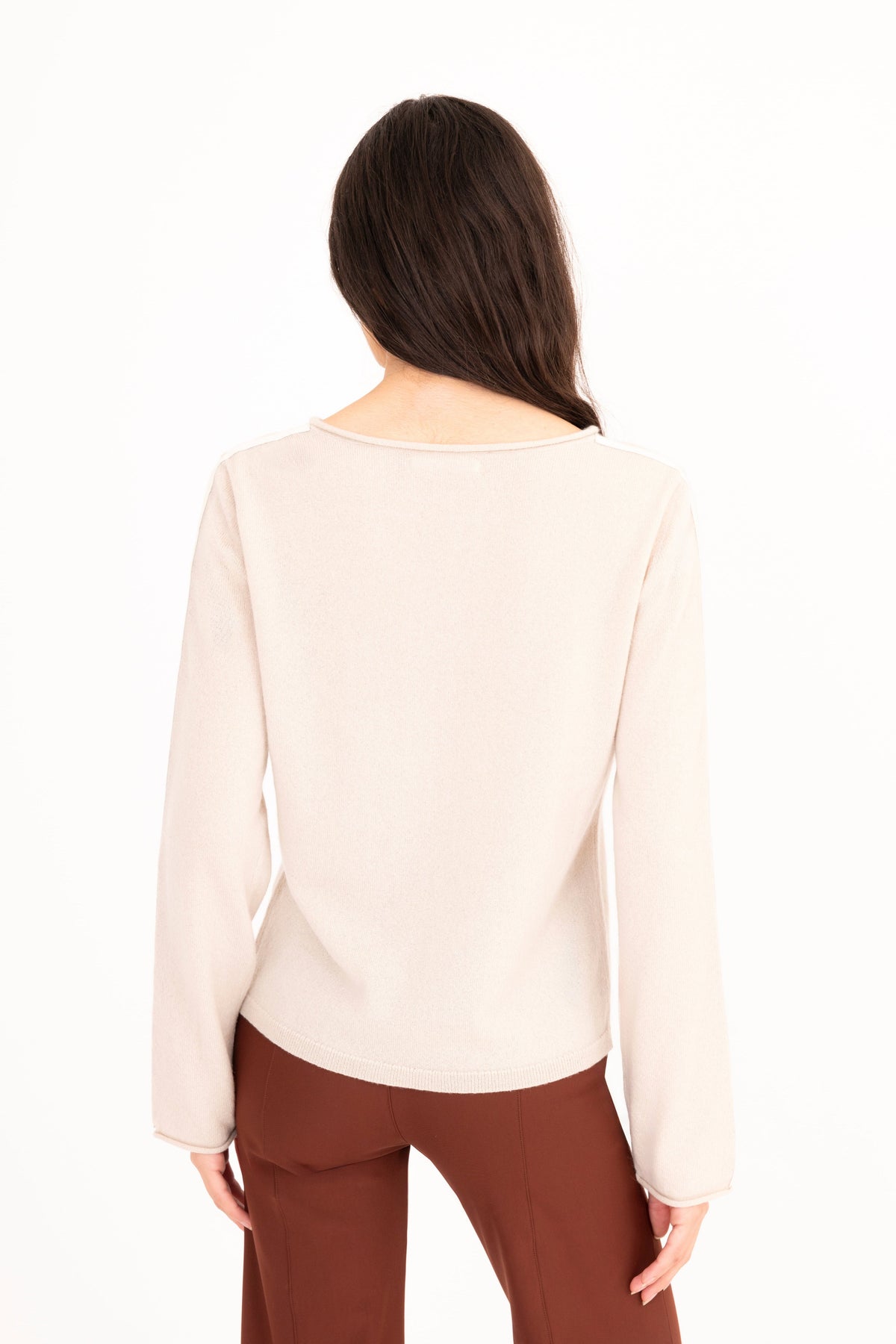 A woman modeling a beige long-sleeve knit sweater, paired with brown pants, against a plain white studio background.