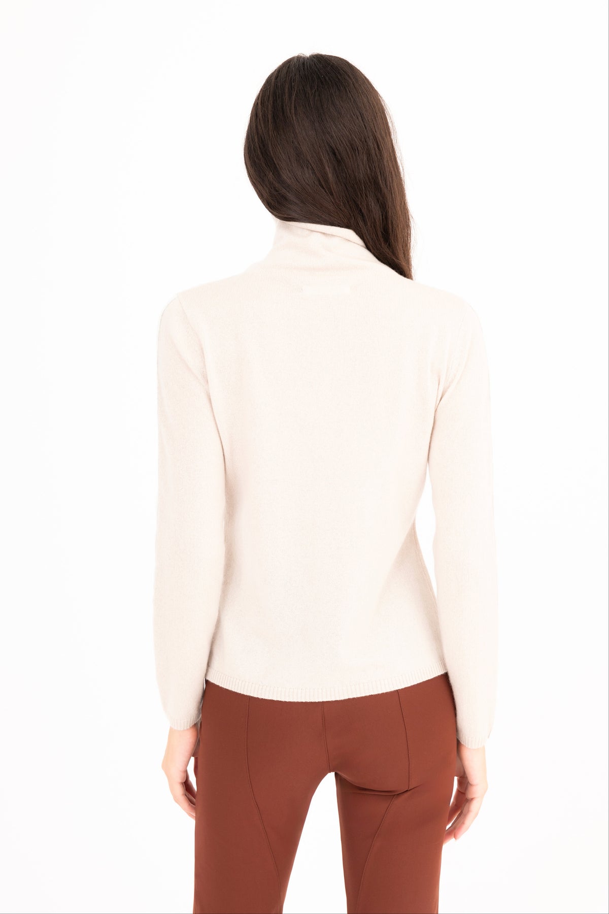 A woman viewed from the back wearing beige long-sleeve mock neck top and brown pants, set against a plain white background.