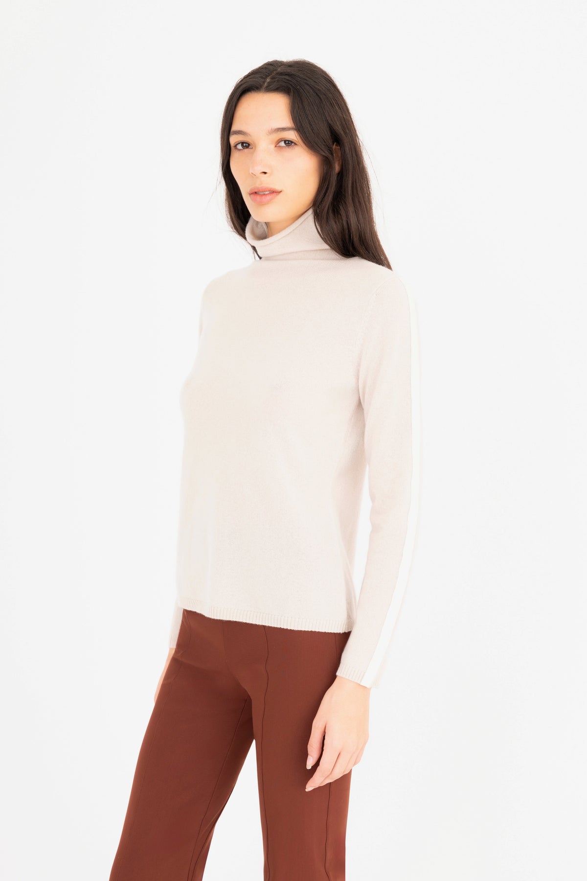 A woman modeling a beige  long-sleeve turtleneck sweater, paired with brown trousers, against a plain white studio background.