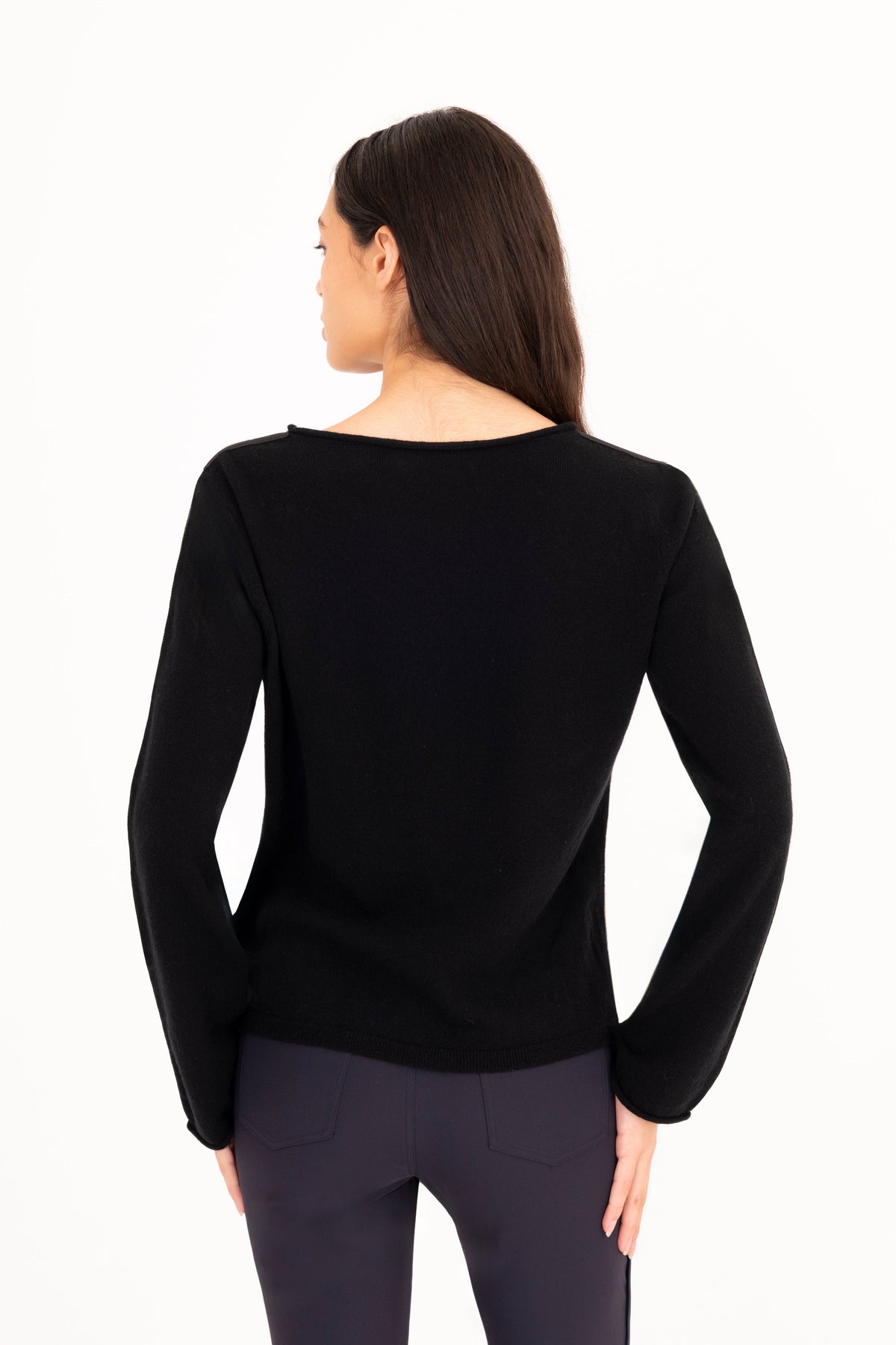 A woman viewed from the back wearing a black long-sleeve knit top and dark navy pants against a plain white studio background.