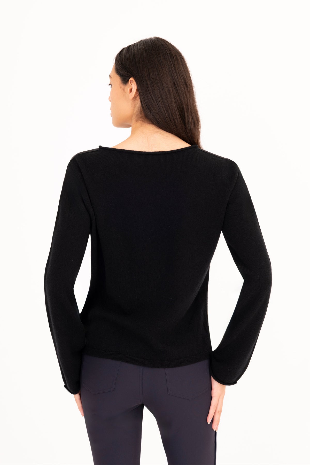 A woman viewed from the back wearing a black long-sleeve knit top and dark navy pants against a plain white studio background.
