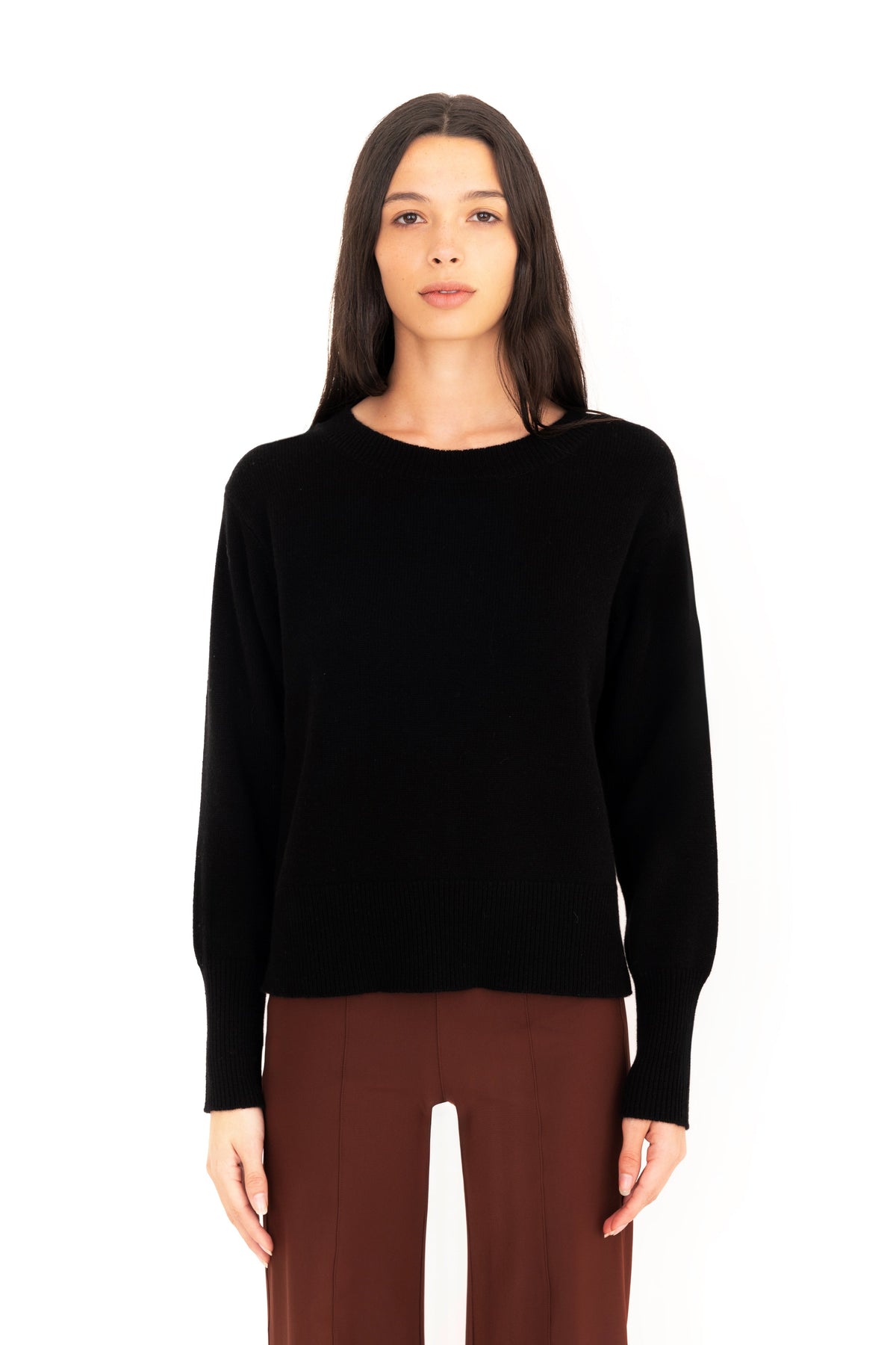 Woman wearing a black crew-neck sweater and dark brown pants on a white studio background.