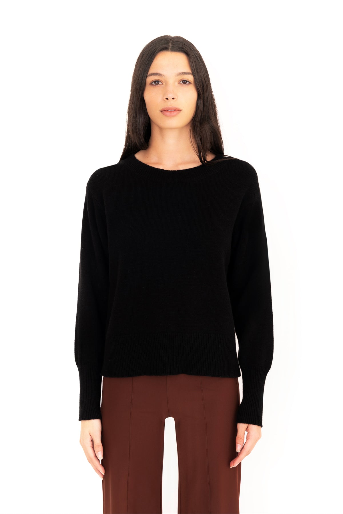 Woman wearing a black crew-neck sweater and dark brown pants on a white studio background.