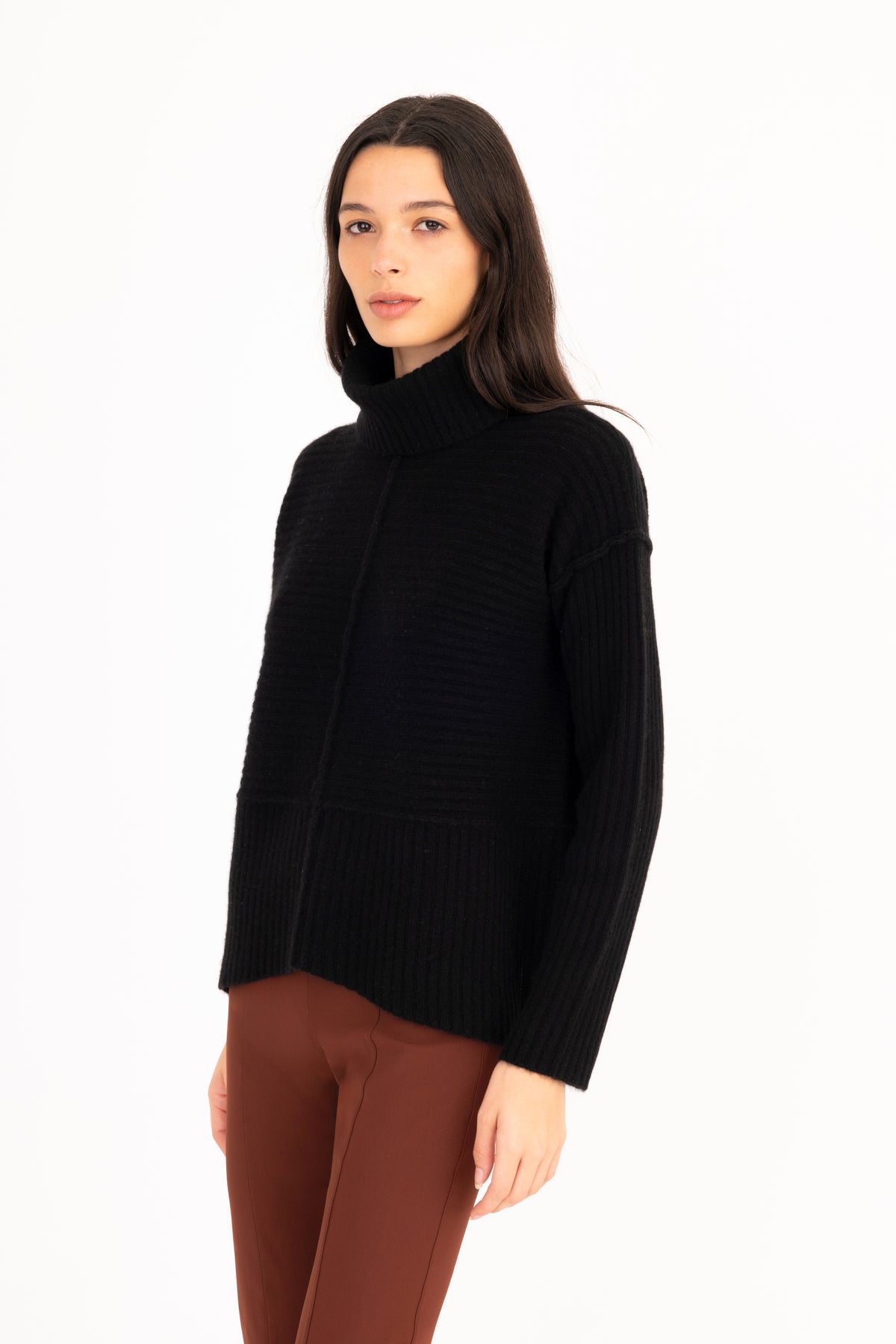 A woman modeling a black ribbed knit funnel-neck sweater with long sleeves, paired with brown pants, against a plain white studio background.