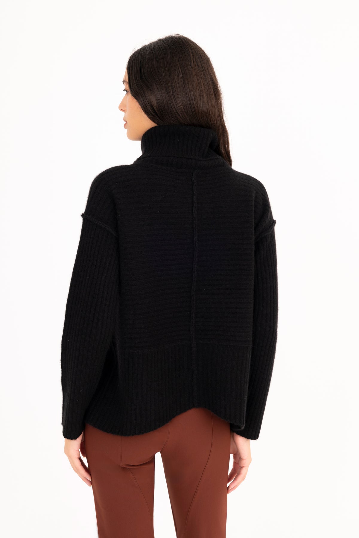 A woman shown from the back wearing a black ribbed funnel-neck sweater and brown pants against a plain white studio background.