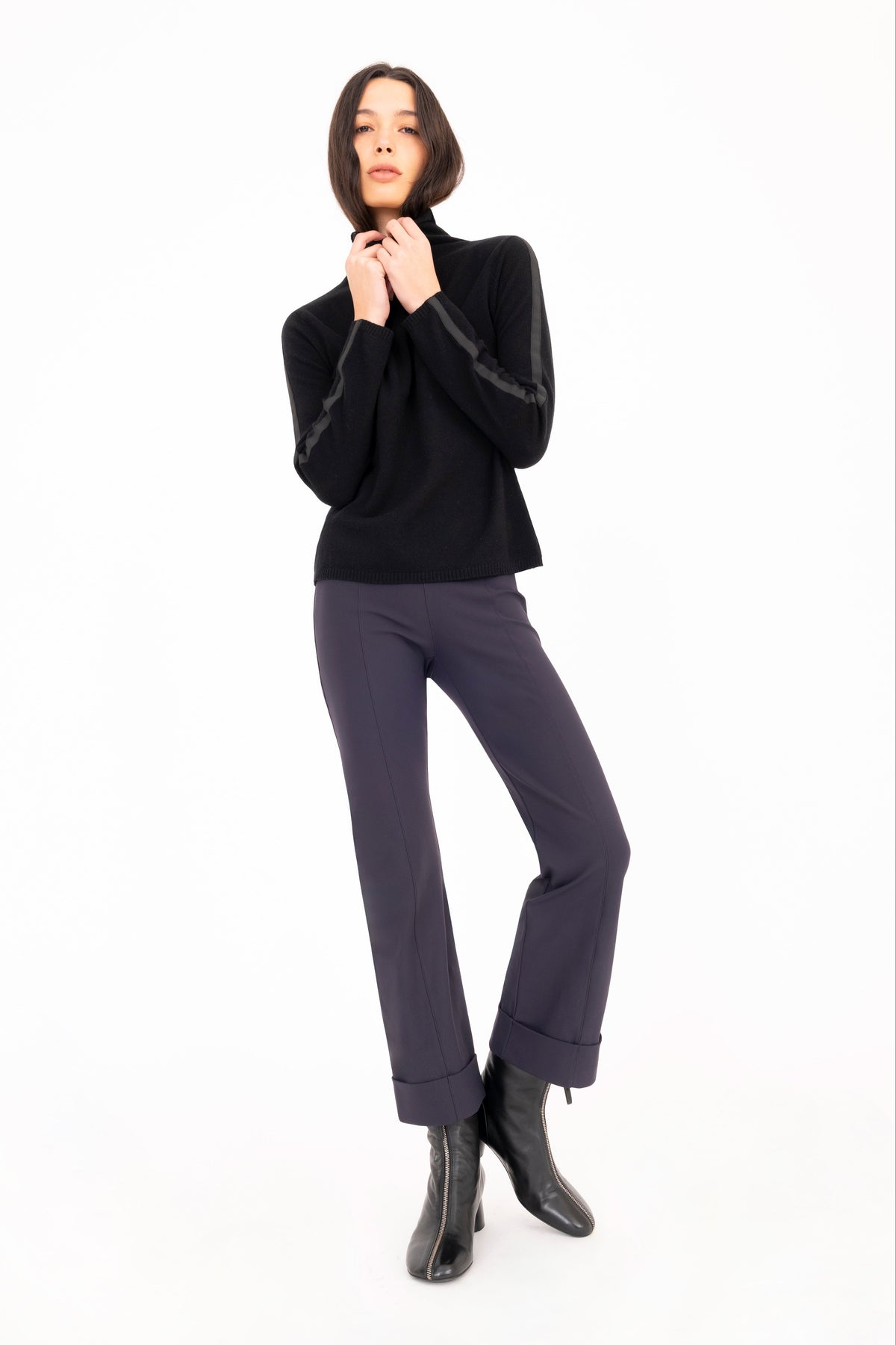 A woman modeling a black long-sleeve mock neck knit sweater, paired with dark navy pants against a plain white studio background.
