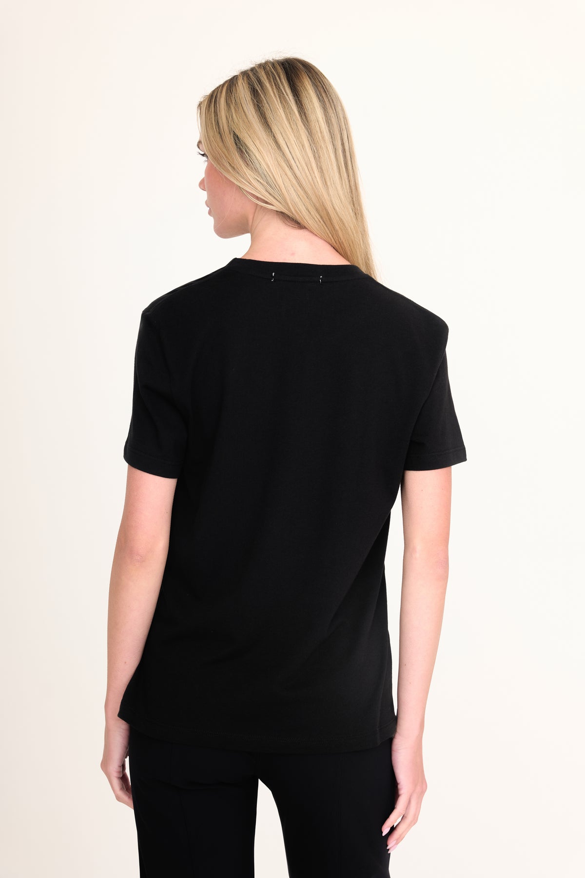 A woman with blonde hair is shown from the back wearing a black short-sleeve crewneck t-shirt and black pants against a plain off-white background.