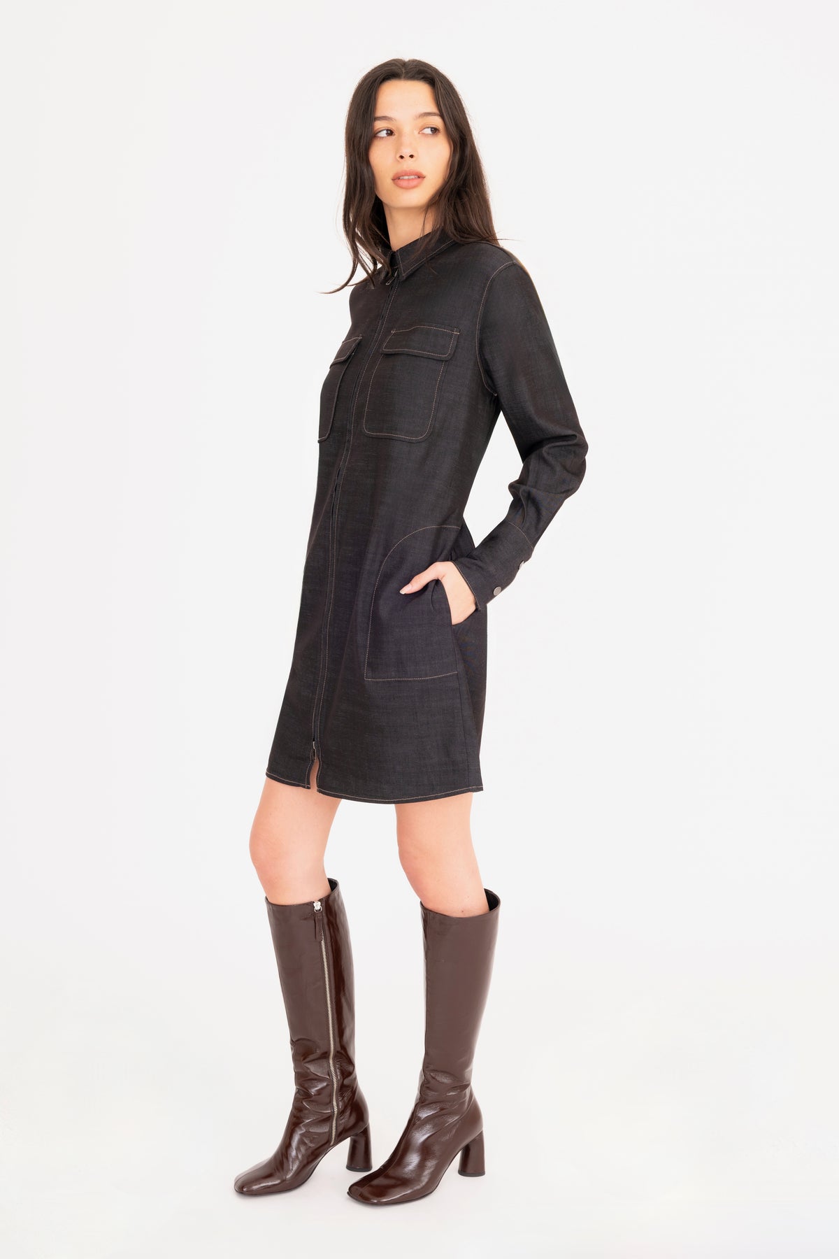 A woman modeling a black denim shirt dress with front pockets, paired with dark brown knee-high leather boots, against a plain white studio background.