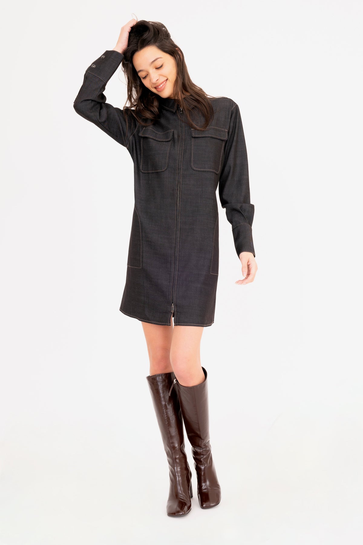 A woman modeling a black denim shirt dress with front pockets, paired with dark brown knee-high leather boots, against a plain white studio background.