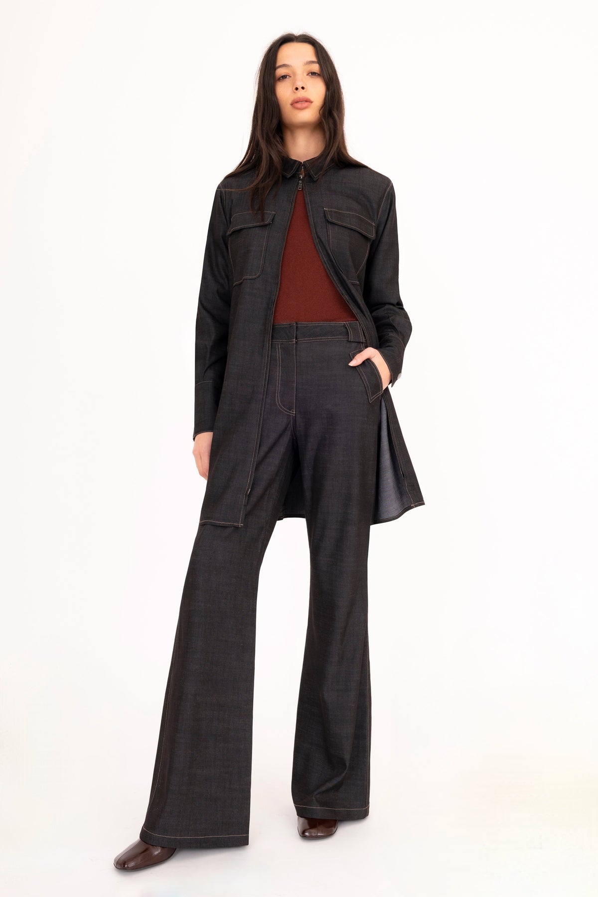 A woman modeling a black denim shirt dress worn as a jacket with matching flared denim trousers, and a brown cashmere crew-neck sweater, paired with dark brown leather boots, against a plain white studio background.