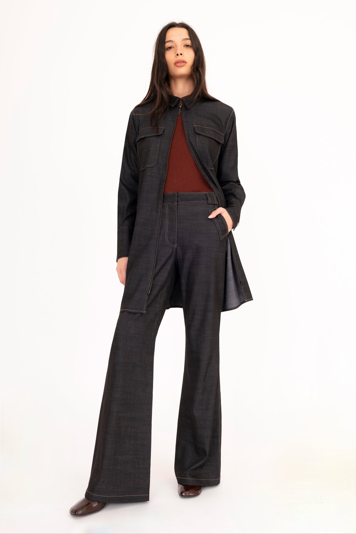 A woman modeling a black denim shirt dress worn as a jacket with matching flared denim trousers, and a brown cashmere crew-neck sweater, paired with dark brown leather boots, against a plain white studio background.