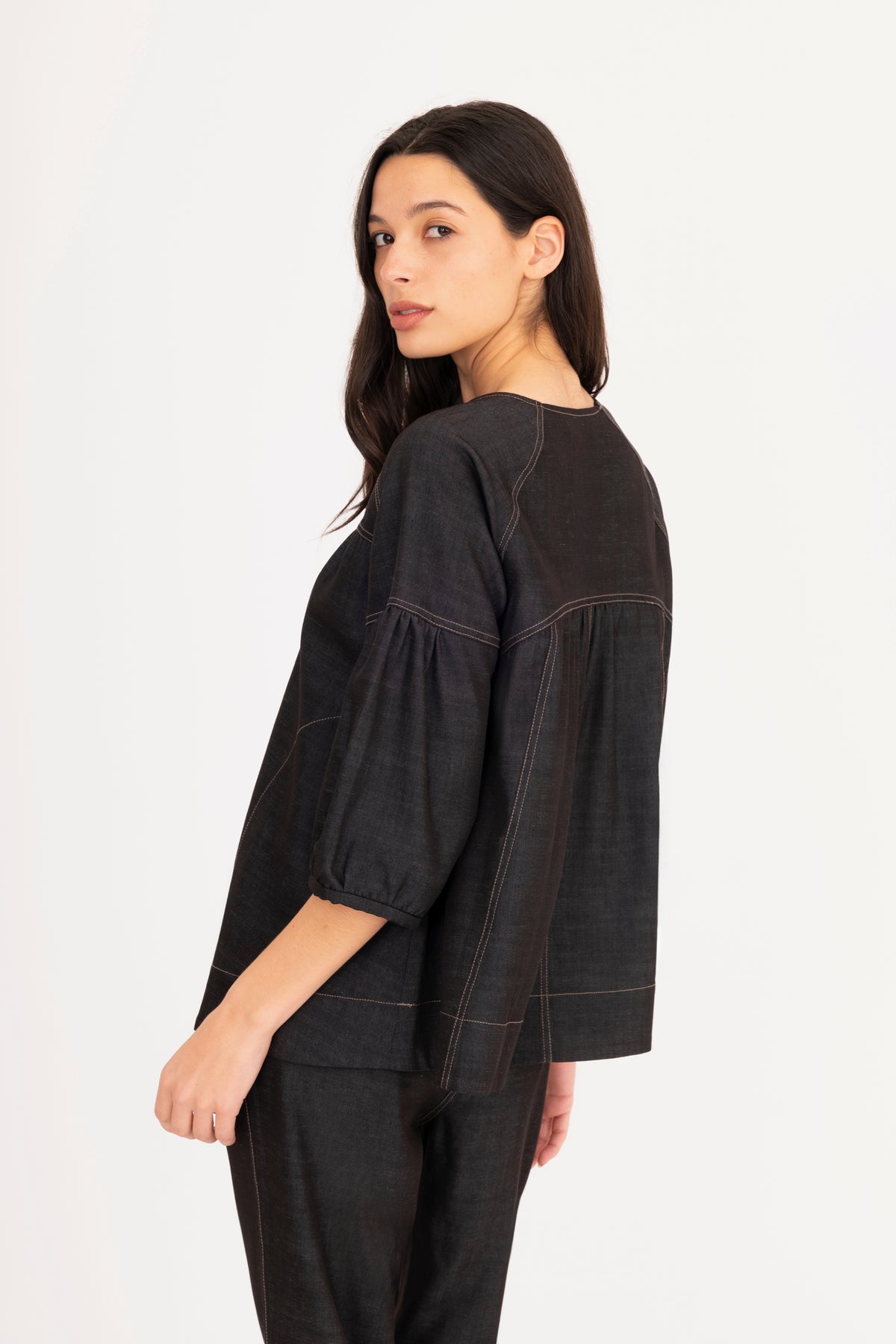 A woman modeling a black denim top with a split neckline, gathered shoulders, and puff sleeves, paired with matching denim pants against a plain white studio background.
