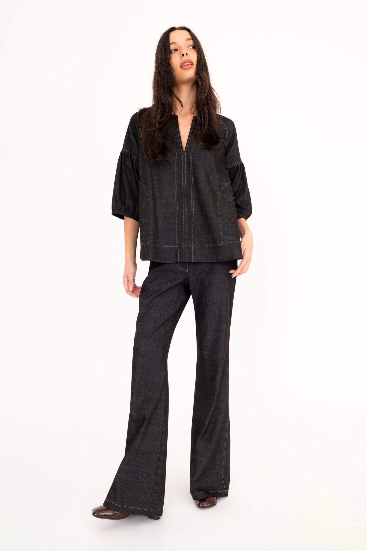 A woman modeling a black denim top with vertical seam details, paired with black denim flare pants and brown boots, against a plain white studio background.
