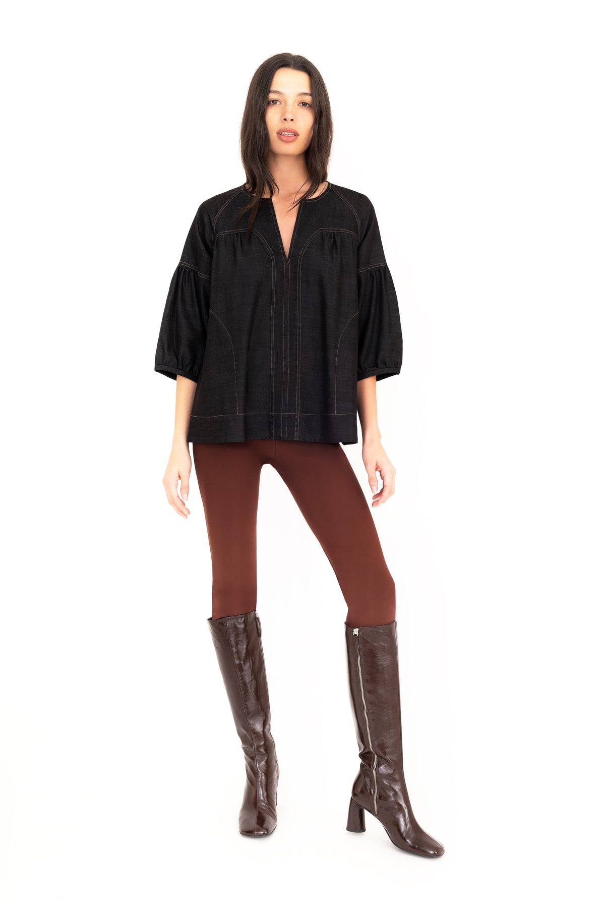 A woman modeling a black 3/4-sleeve top with vertical seam details, paired with brown pants and brown knee-high boots, against a plain white studio background.