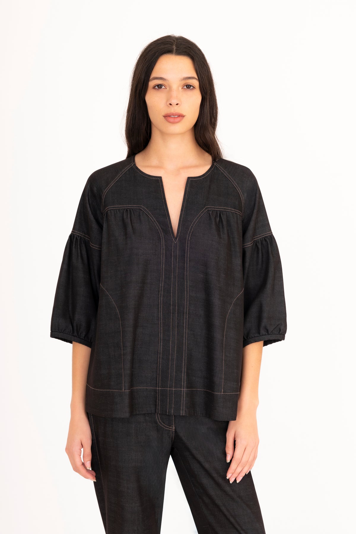 A woman modeling a black denim top with a split neckline, gathered shoulders, and puff sleeves, paired with matching denim pants against a plain white studio background.