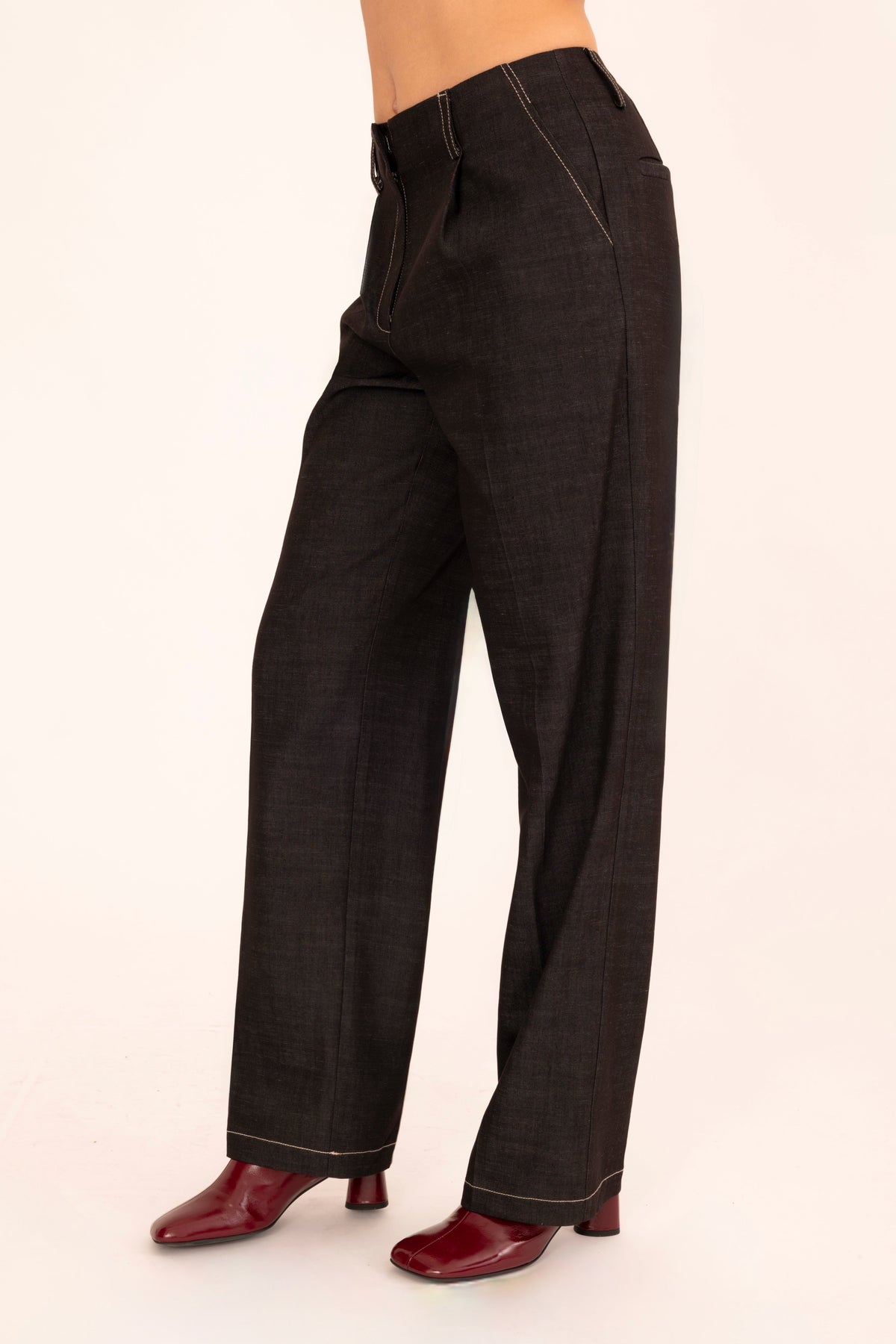 A model is shown from the waist down wearing black denim trousers and dark red shoes, standing against a plain off-white background.